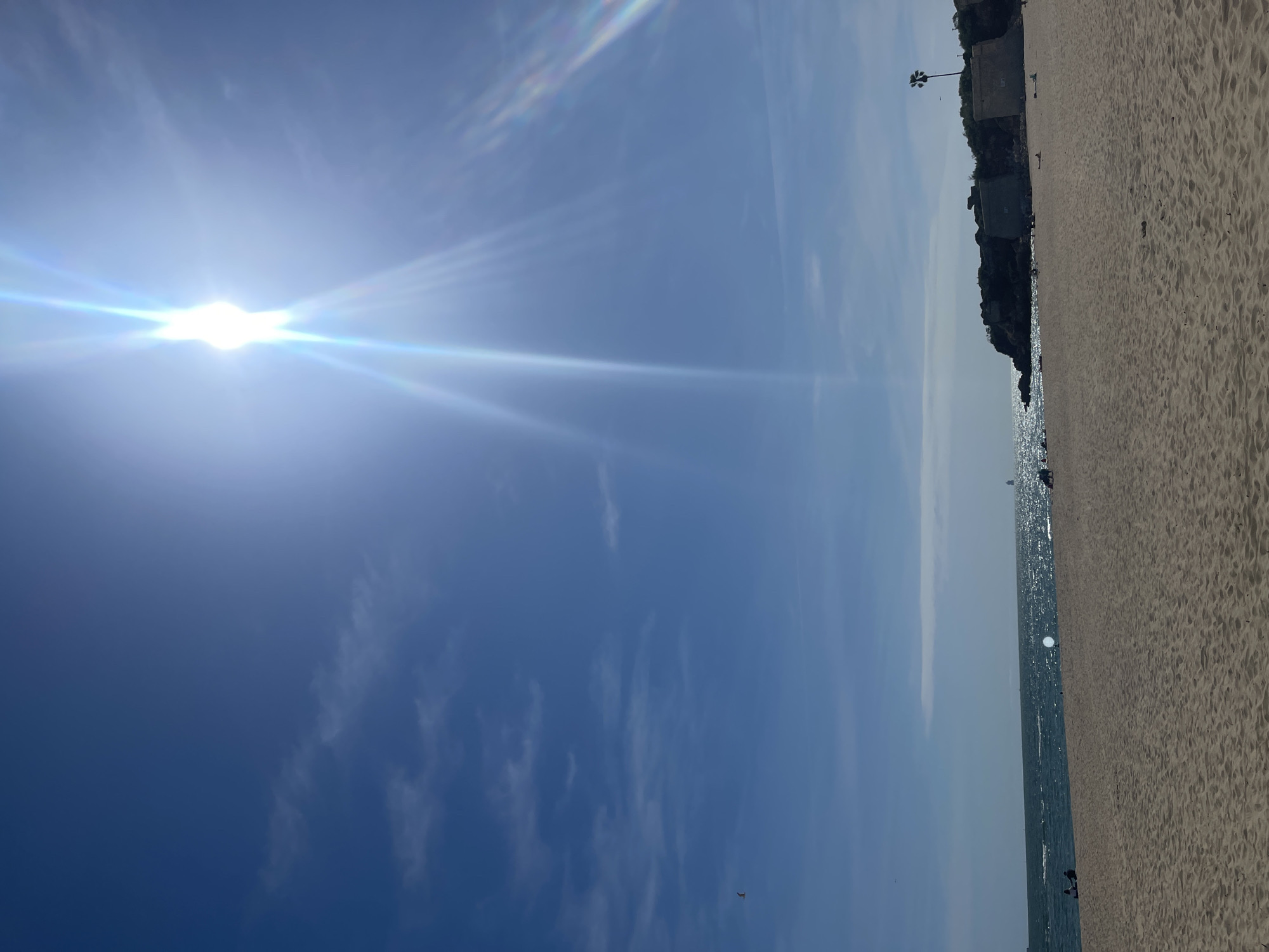 The image shows a beach scene in El Puerto de Santa María, Spain. The bright sun is high in the blue sky, causing a glare. The beach, a sandy expanse, occupies the right side of the frame. The sea is a narrow strip with sparkling water. In the background, there is a cliff edge covered in greenery, and a palm tree is visible. People can be seen on the beach. The picture is taken in a way that the horizon is vertically oriented.