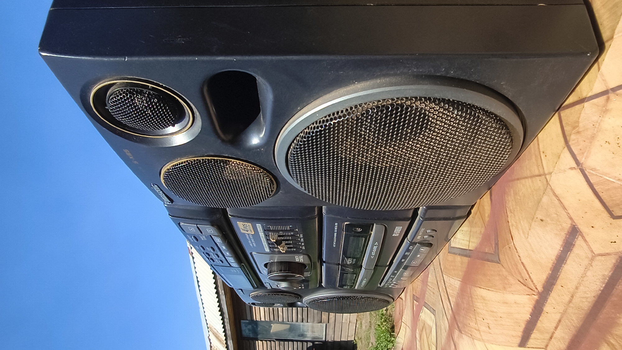 A black Panasonic boombox sits on a patterned, light brown surface outdoors, with a bright blue sky in the background and a wooden building with a corrugated metal roof to the left. The boombox has several speakers, a cassette deck, and various buttons and controls. The surface it rests on appears to be a tarp or mat with a geometric design. This scene is located in Mikhaylovka, Russia.