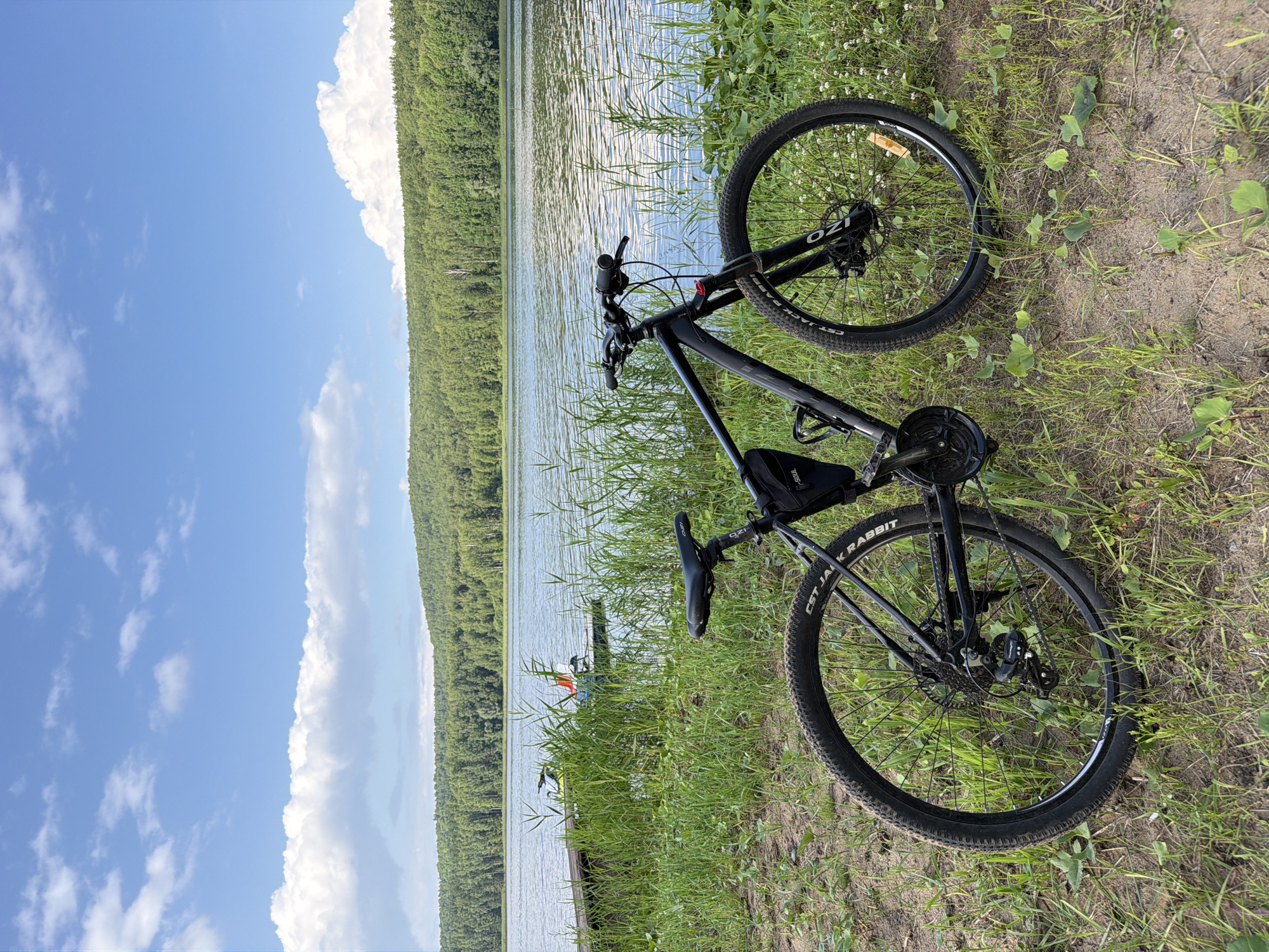 A black mountain bike is parked on a grassy area next to a calm body of water. The bike is positioned in the foreground, facing towards the right side of the frame. It has a saddle bag attached to the seat post and a water bottle cage on the frame. The rear wheel is clearly visible, with the brand name "CST JACKRABBIT" printed on the tire. The front of the bike, including the handlebars and front wheel with disc brakes, is also visible.
In the middle ground, the water stretches across the frame, with gentle ripples on its surface. A wooden dock extends into the water from the left side, with two small boats or jet skis moored to it. The boats appear to be light-colored with some red accents. Tall green grass and reeds grow along the shoreline, partially obscuring the view of the boats.
The background features a dense, green forest that covers rolling hills. The trees are a vibrant green, indicating a warm season. Above the trees, a bright blue sky is dotted with white, fluffy clouds. The overall impression is a serene natural landscape, likely a park or recreational area. While the specific location in Chaykovsky, Russia is not visually identifiable, the scene depicts a common natural setting found in such regions. There are no people present in the image.