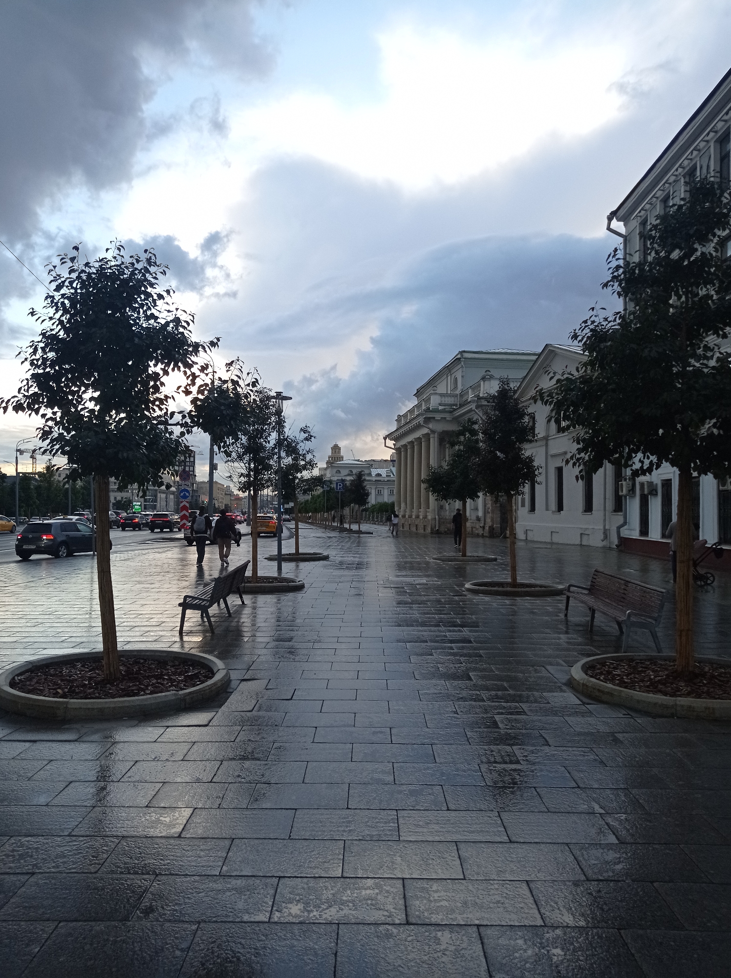 The image depicts a street scene in Moscow, Russia, likely on a cloudy or post-rainy day. The foreground is dominated by a wet, paved walkway made of dark, rectangular tiles, reflecting the dim light. Several young trees in circular planters are spaced along the walkway, some with wooden benches beside them. In the middle ground, a street with moderate car traffic is visible, with pedestrians walking on the sidewalk. On the right side of the street, a large, classical-style building with columns and a white facade stands, typical of Moscow's historical architecture. The sky is overcast with dark clouds, suggesting the possibility of recent or imminent rain.