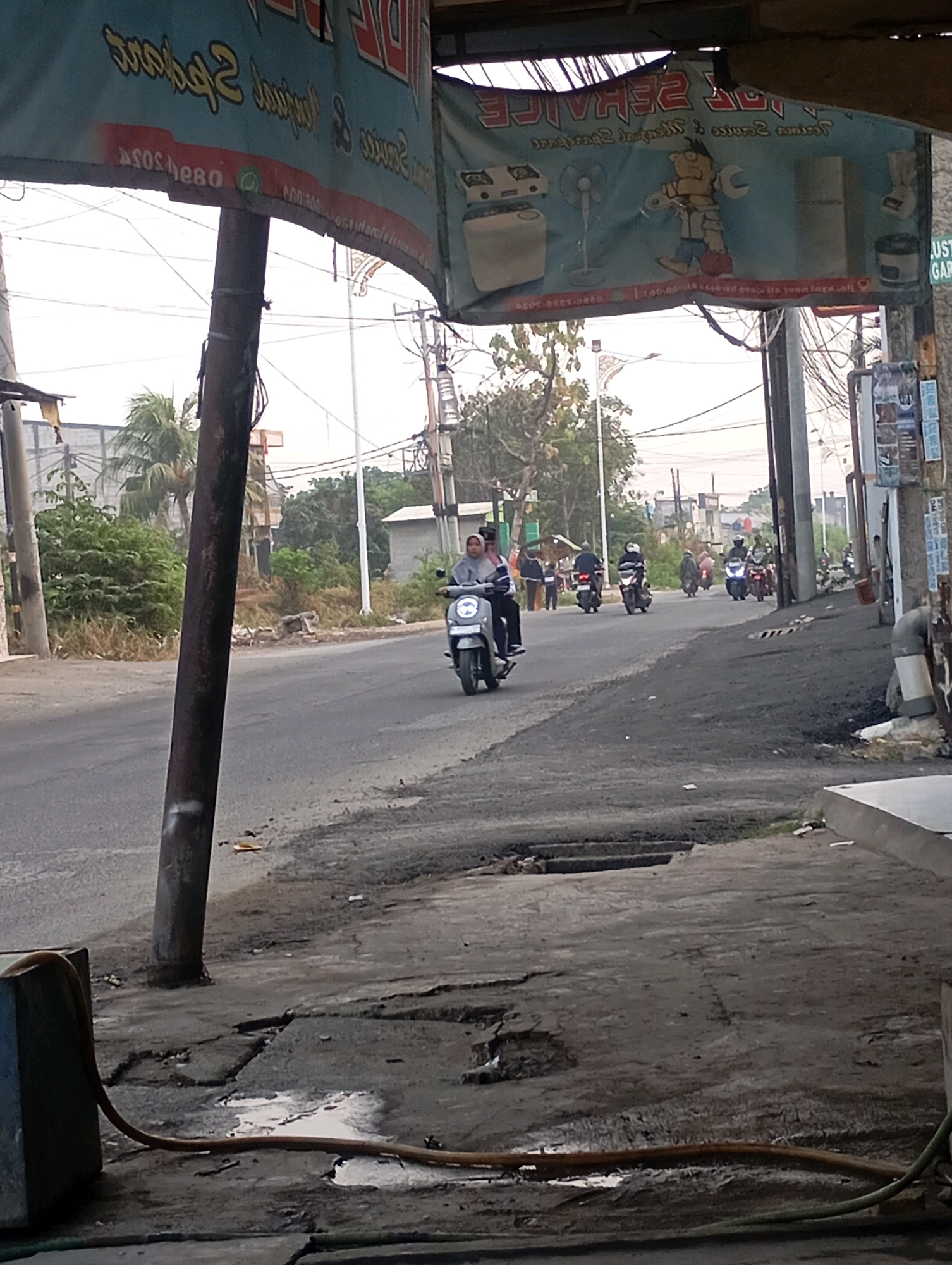 In Babelan, Indonesia, a street scene is captured. A banner advertising "Service" and showing cartoon characters is visible, partially obscured by a pole. The banner contains text that appears to be a phone number and possibly a WhatsApp icon. Several motorcycles are on the road, with some riders visible. One motorcycle in the foreground is ridden by a person wearing a hijab. The road appears to be asphalt with some cracks and patches of darker material. On the right side of the image, a pole with various notices is present, along with a pipe. The background shows buildings and trees, indicating a semi-urban environment. The lighting suggests it is daytime.