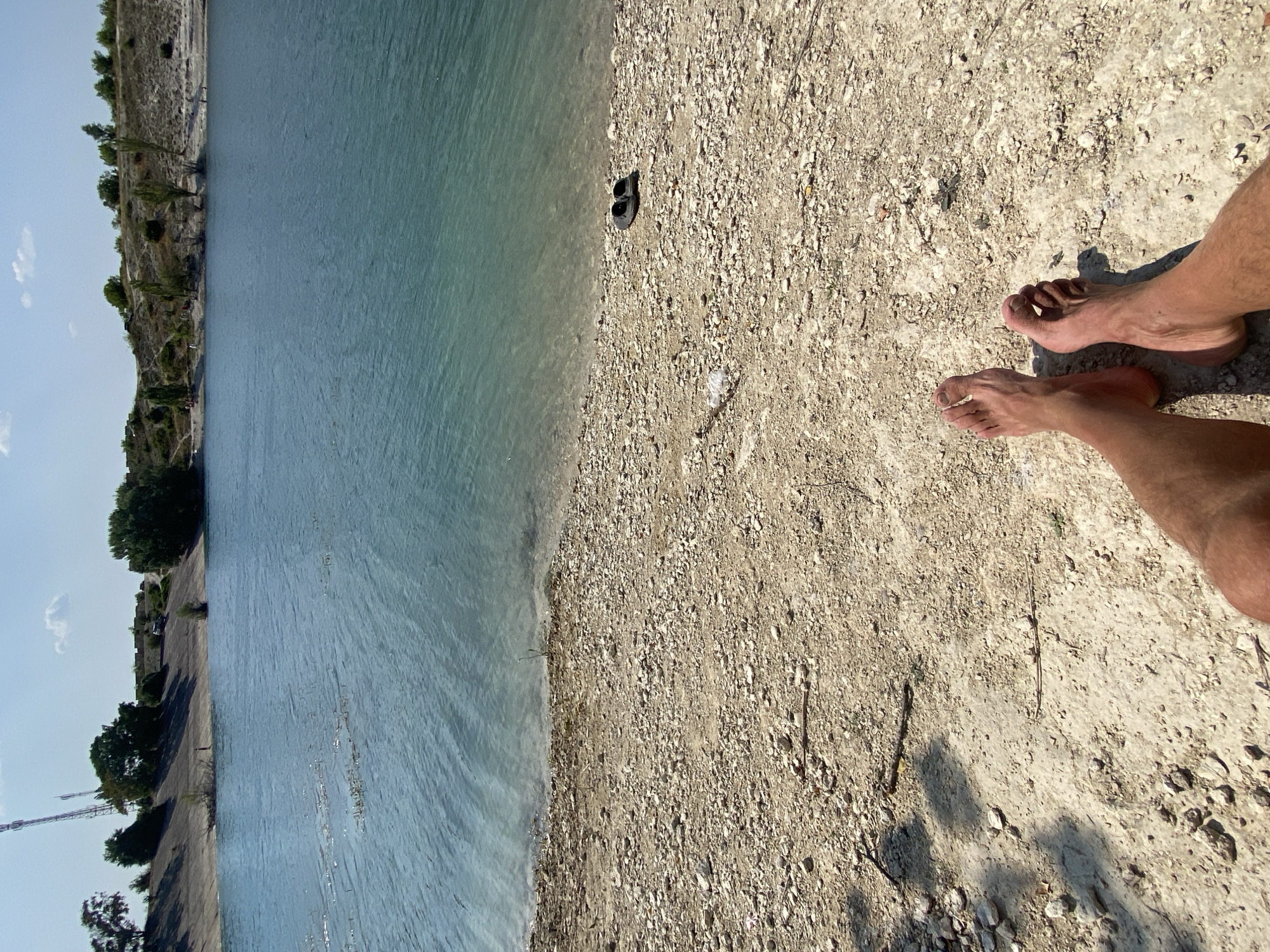 The image shows a view of a body of water, likely a reservoir or lake, with a shoreline of pebbles and sand in the foreground. Two bare feet are visible in the bottom of the frame, resting on the pebbled ground. A pair of dark-colored flip-flops are placed on the shoreline further back from the feet. The water is a clear turquoise color, and the far bank of the water is lined with trees and gently sloping hills under a bright, clear sky with a few scattered clouds. The overall scene suggests a recreational area, possibly a beach or a place for swimming and relaxation. The location is identified as the city of Куйбышевское сельское поселение, Ukraine.
