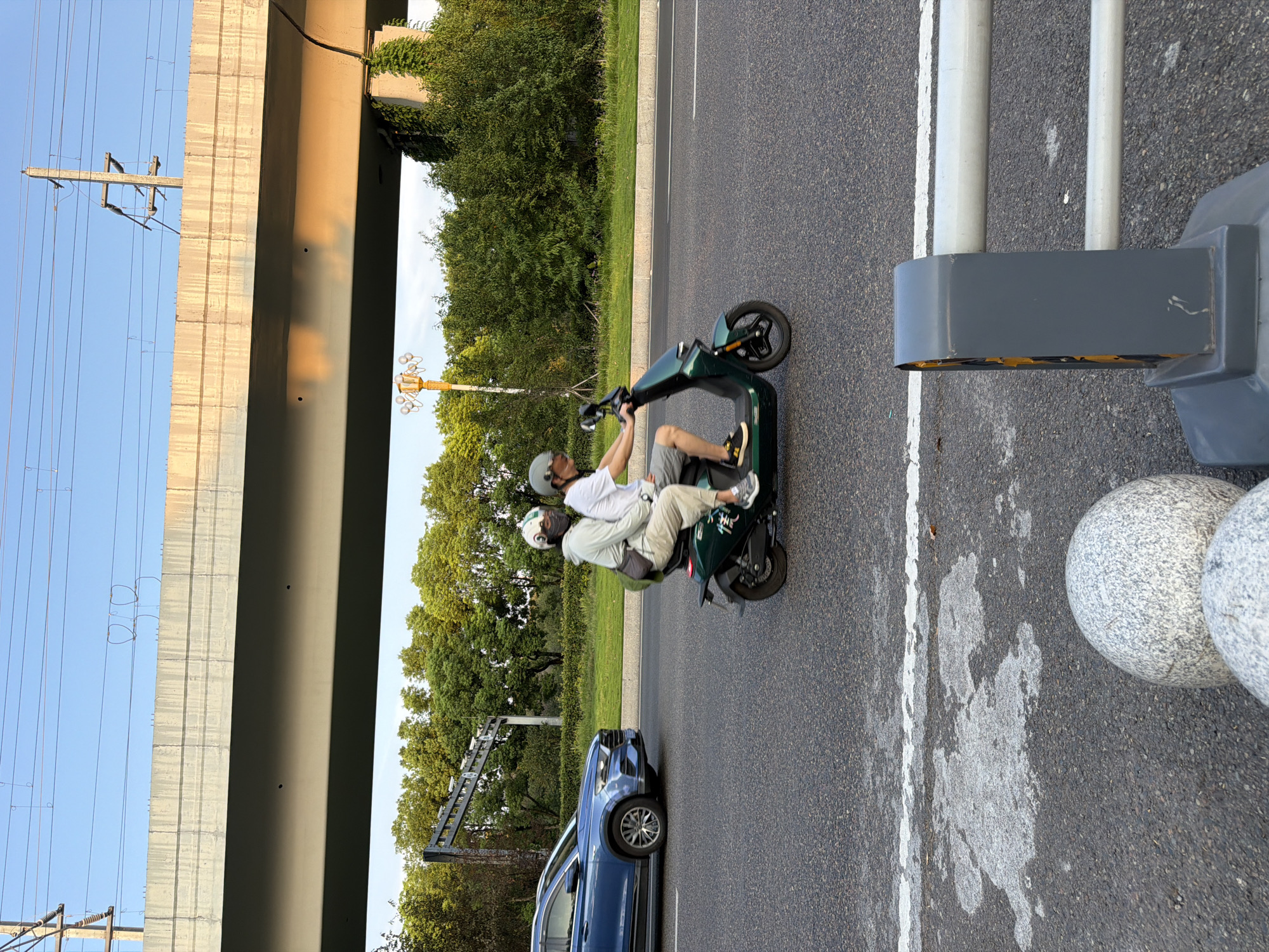 Two individuals, wearing helmets, ride a green electric scooter on a paved road. The person in front is wearing a light-colored long-sleeved shirt and light-colored pants, and has a dark-colored bag slung across their shoulder. The person in the back is wearing a white short-sleeved shirt and grey shorts. They are positioned under an elevated concrete structure with overhead power lines. To the left, a blue SUV is driving on the same road. The background consists of dense green trees. The scene appears to be in the city of Jiangjin, China, given the context.