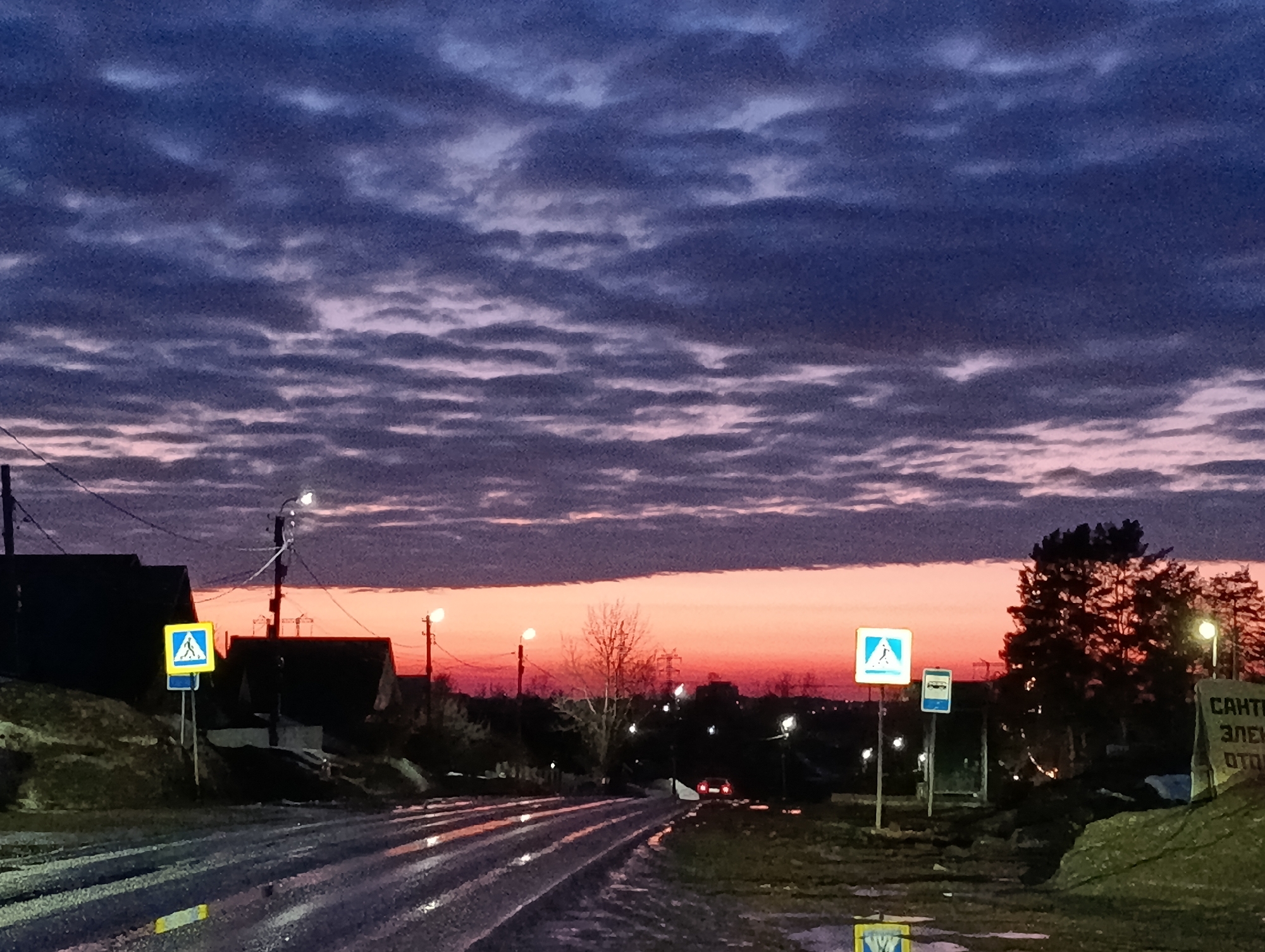 The image depicts an urban street scene at twilight in Chaikovsky, Russia, characterized by a deep blue-purple sky with scattered altocumulus clouds transitioning to a vivid orange-red horizon. A wet, paved road, reflecting ambient light and street illumination, curves gently from the foreground into the distance.
On the left side of the road, dark-silhouetted buildings with sloped roofs are visible on an elevated terrain. A utility pole supports power lines and two active streetlights, casting light onto the immediate area. A yellow-and-blue square pedestrian crossing sign (GOST 52289-2004 type 5.19.1) is prominently positioned beside the road.
In the middle ground, additional illuminated streetlights line the road, and a bare deciduous tree stands against the colorful horizon. A single vehicle, identified by its illuminated red taillights, is observed receding in the distance.
The right side of the road features another yellow-and-blue pedestrian crossing sign, a smaller blue rectangular sign with an indiscernible white graphic, and a cluster of dark coniferous trees. A large, rough-textured stone or concrete slab on the far right displays partially legible Cyrillic text: "САНТЕХ ЭЛЕКТРО ОТОПЛЕНИЕ" (Plumbing, Electrical, Heating). No human figures are present in the scene.
