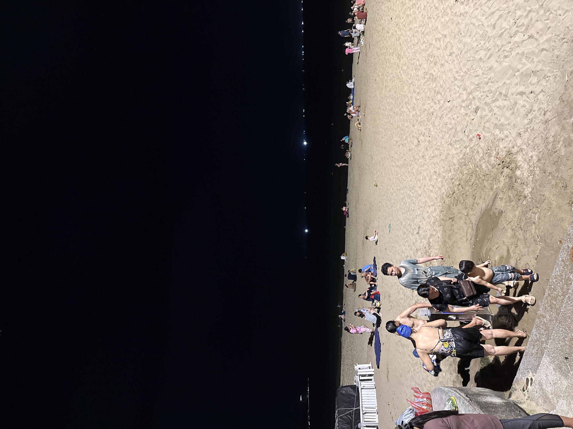 This image shows a nighttime scene at a beach in Nha Trang, Vietnam. The sky is dark, with a thin sliver of lights visible on the horizon, suggesting a populated coastline. The sand is a light tan color, with footprints and ripples indicating recent activity.
In the foreground, a group of people are gathered near a concrete embankment and stairs. A woman wearing a patterned swimsuit and black shorts, with a blue cap, is turning away from the camera. She appears to be holding a towel. Beside her, a woman in a black floral dress is walking down the stairs, carrying a brown purse. A young boy in striped shorts is walking down the stairs in front of her. Behind them, a man in a grey t-shirt stands looking to the side. Further back, more people are visible on the beach, some sitting on the sand, others walking.
In the mid-ground, a cluster of people are spread out on the sand. Some are sitting together on blankets, while others are standing or walking. There are white structures on the left side of the image, possibly part of a beachside establishment or temporary shelter. The ocean is visible in the background, appearing dark under the night sky, with faint lights reflecting on its surface. The overall atmosphere suggests a casual evening gathering on the beach.