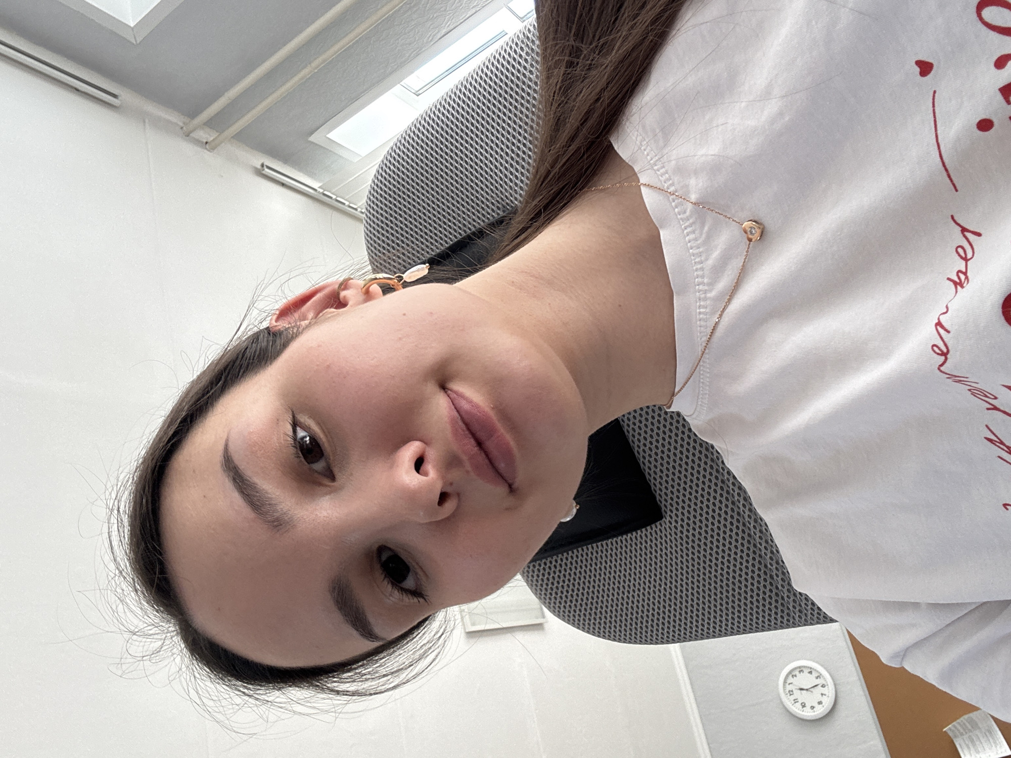The image shows a young woman seated in a gray mesh office chair. She is wearing a white long-sleeved shirt with red text on it. She is wearing a gold necklace. Her location in the city of Chaikovsky, Russia is not determinable from this image.