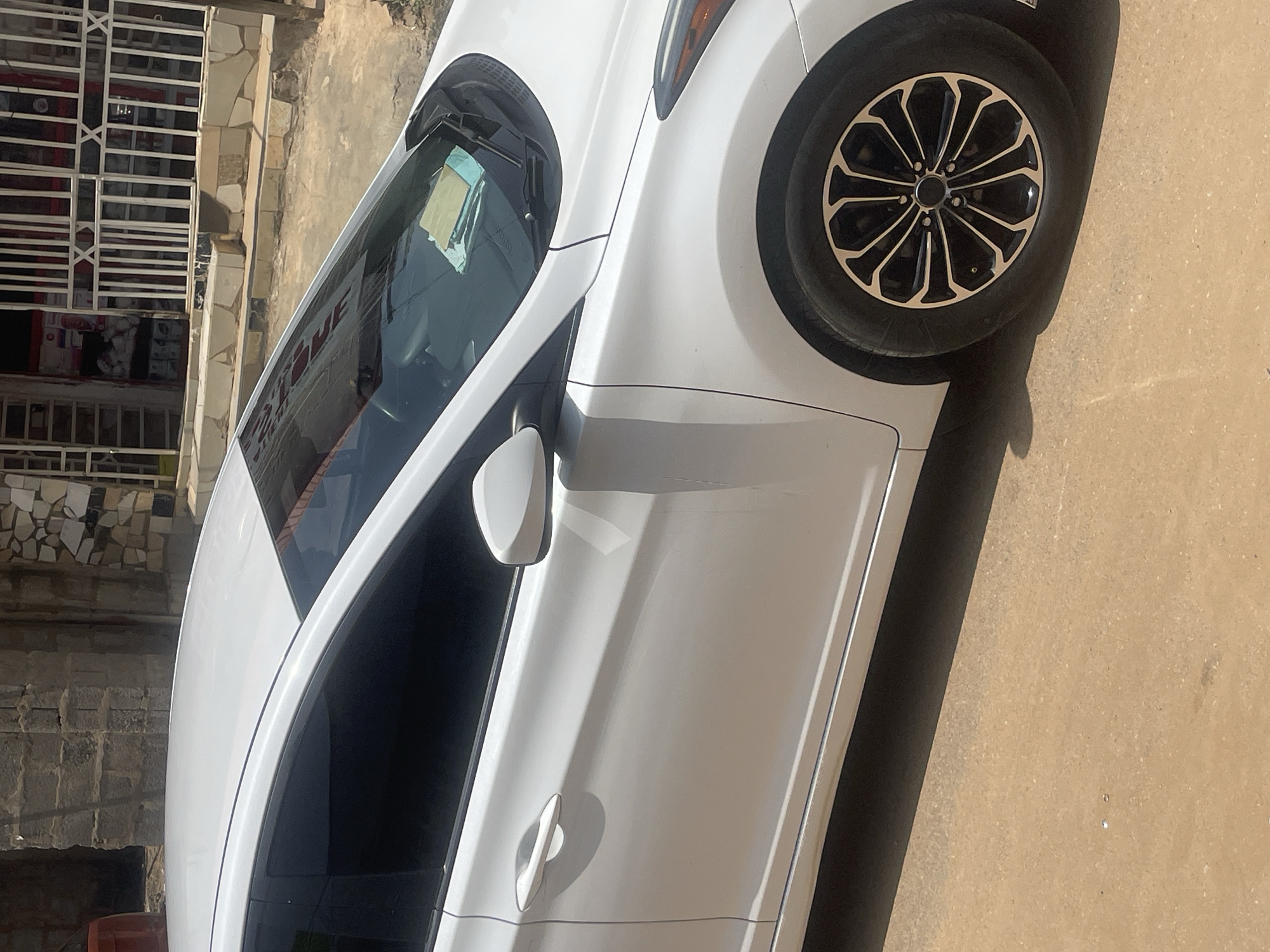 A white sedan is parked on a dirt road in front of a building with a white metal fence. The car's driver-side door and front wheel are visible. The building appears to be a shop with goods displayed inside. The location is the city of Abuja, Nigeria.