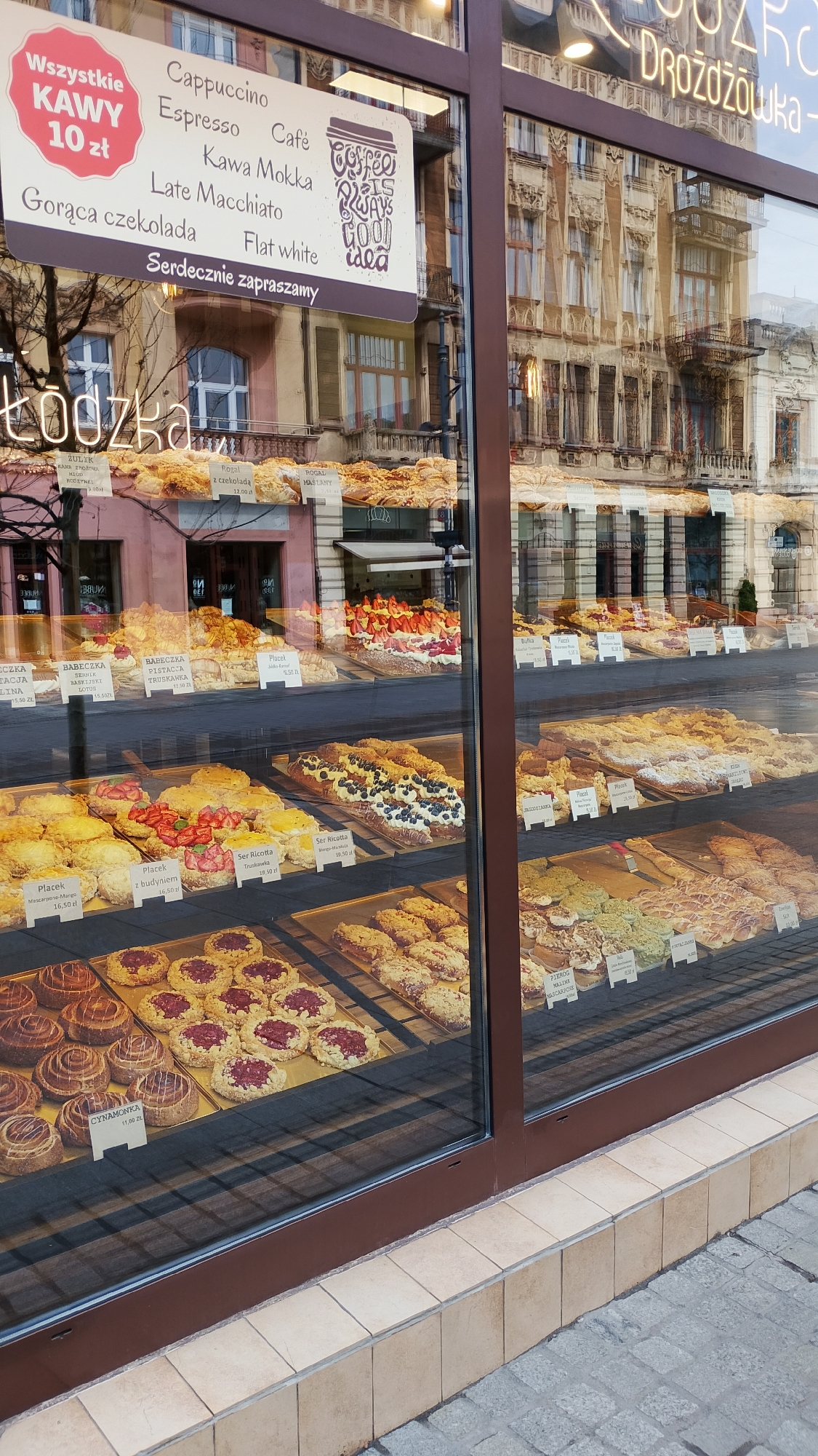 A street-level view of a bakery storefront in Łódź, Poland, showcases a window display filled with an array of pastries. The window reflects the surrounding urban environment, revealing detailed architectural facades of adjacent buildings. A sign advertising coffee and other beverages in Polish is visible, including prices. The various baked goods on display include cinnamon rolls, cakes, and other pastries with fruit and cream fillings. Price tags are displayed next to each pastry. The sidewalk outside is visible in the foreground.