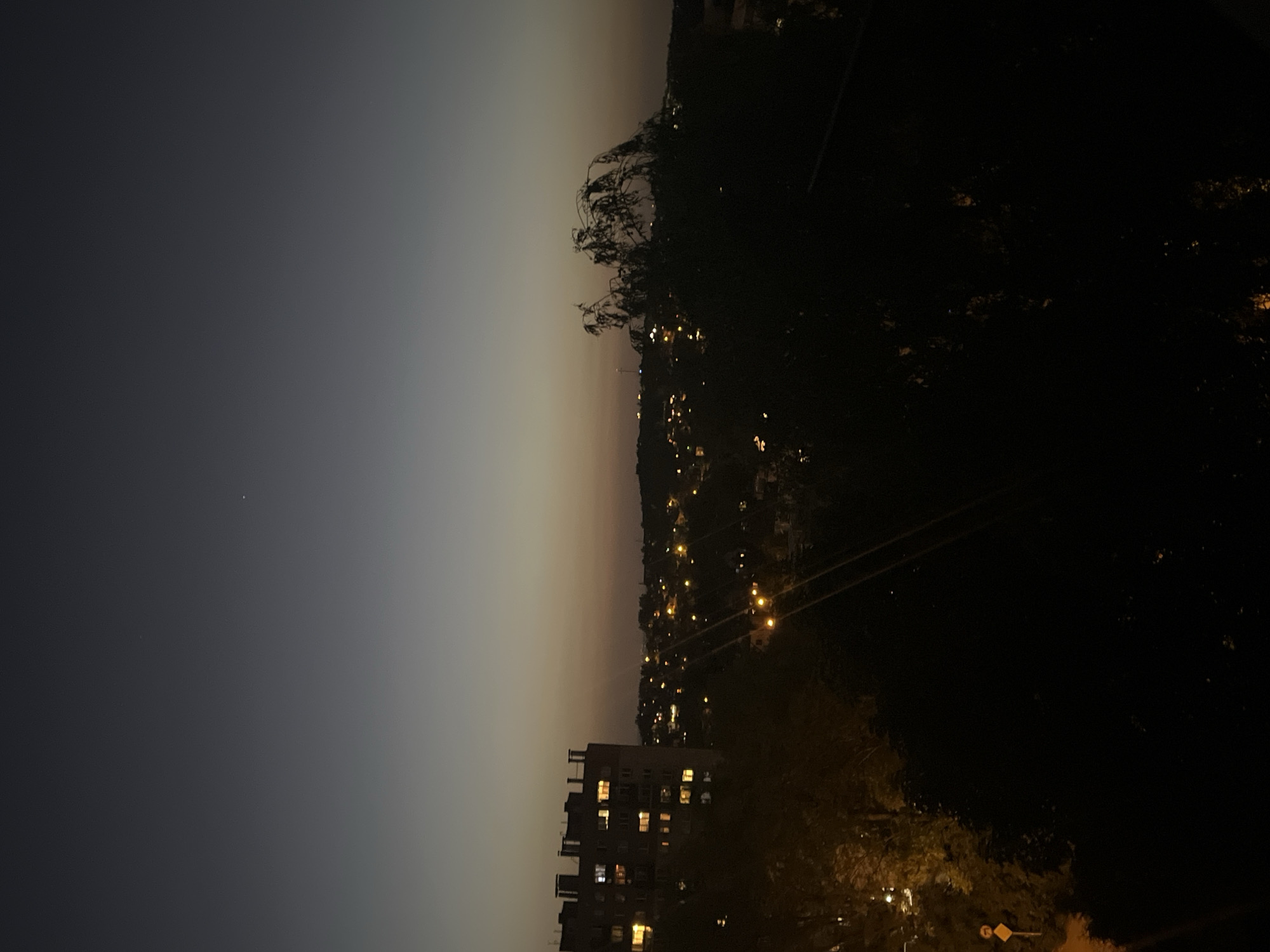 The image shows a nighttime view of a city. The sky transitions from dark gray to a lighter hue near the horizon. A building with lit windows is visible in the foreground, suggesting residential or commercial occupancy. Power lines stretch across the scene. Trees and foliage obscure some of the view, contributing to the darkness. Distant lights indicate the presence of a populated area beyond the immediate foreground.