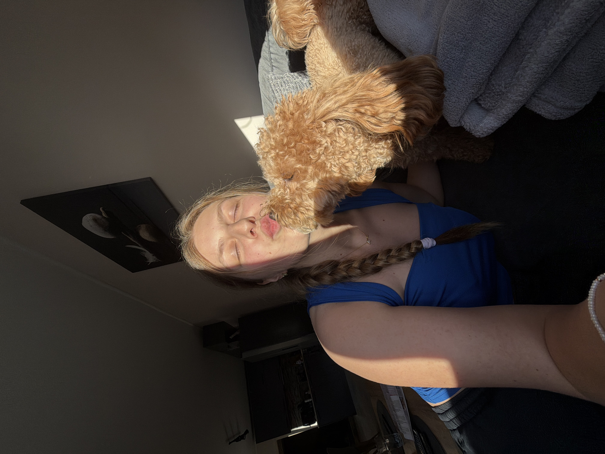 A young woman with blonde hair in a braid, wearing a blue t-shirt, is lying on a dark-colored sofa or couch. She is positioned with her head and torso angled towards the viewer, and her arm extending to the right side of the frame. A medium-sized, light-brown, curly-haired dog is perched on top of her, seemingly kissing her face. A light gray blanket is draped behind the dog on the couch. To the left of the woman, there's a dark wall with a framed black and white picture. A dark cabinet-like structure is visible in the background. The lighting appears to be coming from a window, casting shadows on the wall and woman. The location in Litakovo, Bulgaria, cannot be determined from the image.