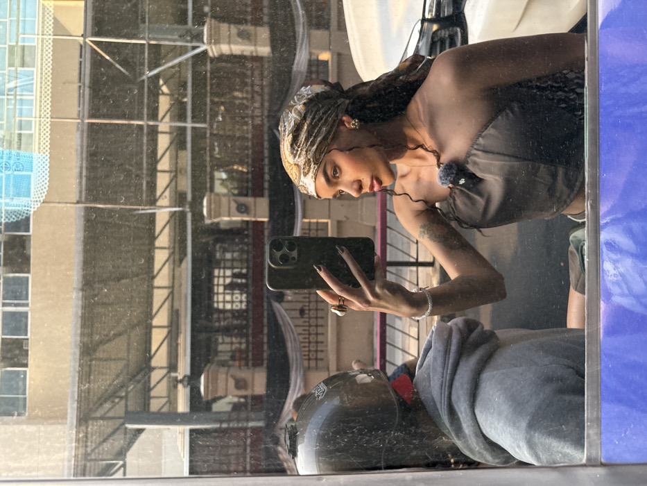 A selfie is captured in a reflective glass surface on an urban street in Bangkok, Thailand. The primary subject is a young woman, visible from the chest up, holding a dark-colored smartphone in her right hand to take the photograph. She wears a patterned headscarf (gold, brown, white), a dark brown one-shoulder top with a fuzzy accessory attached near the neckline, a bracelet, and rings. A tattoo is visible on her right upper arm. To her left, a secondary subject is partially reflected, wearing a dark helmet with a red strap and a grey hooded sweatshirt, suggesting they are associated with a motorcycle or scooter. The background reflection shows an urban street scene featuring a multi-story building with a light-colored facade and windows, a lower structure with a corrugated roof, decorative pillars, and a dark fence. A portion of a light-colored vehicle is visible on the far right. The scene is illuminated by direct sunlight.