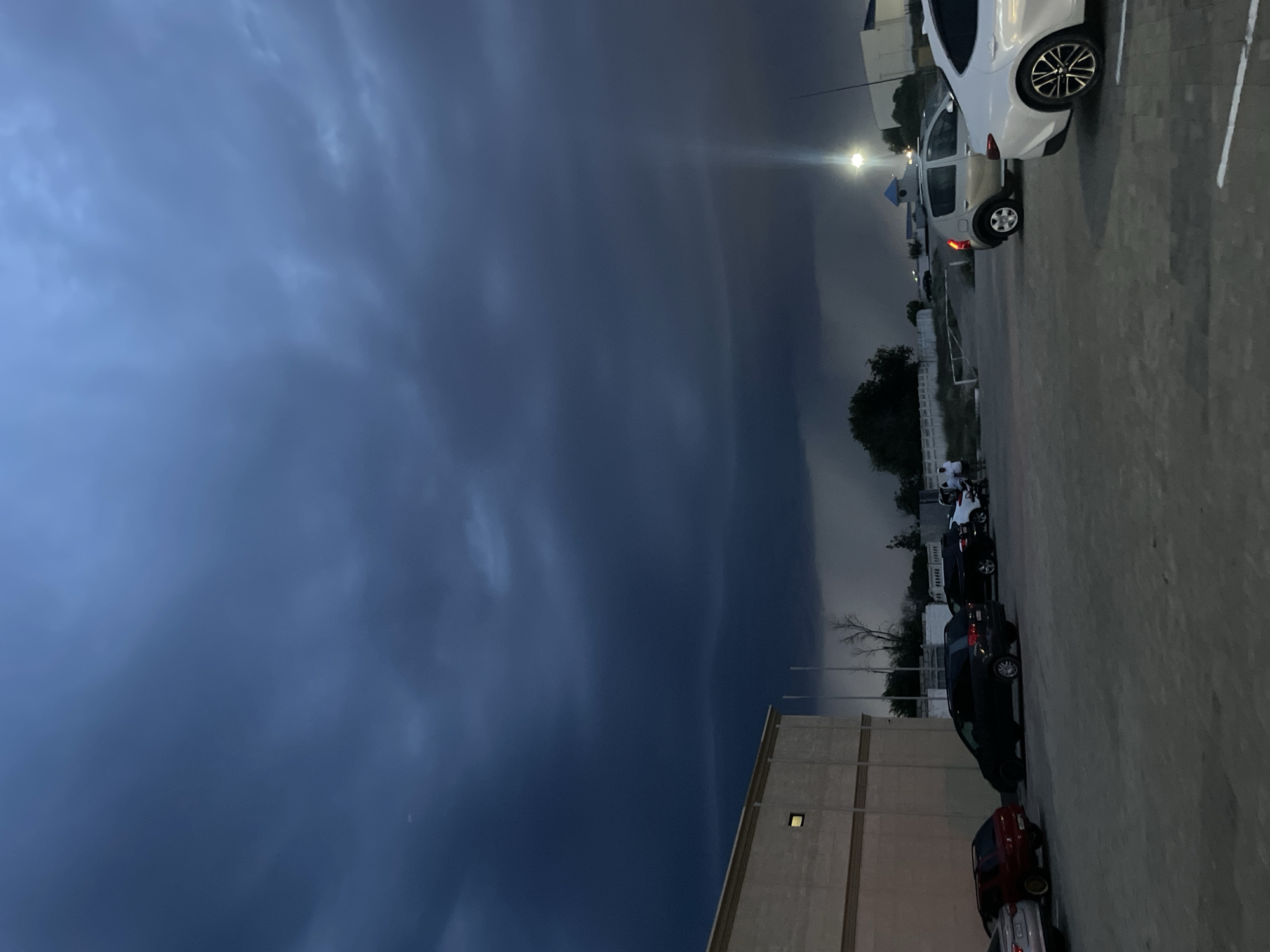 The image shows a parking lot in the city of Aktau, Kazakhstan, under a dark, stormy sky. Several cars are parked in the lot, including a silver SUV, a black SUV, a red hatchback, and a white sedan. A building with a beige facade is visible on the left side of the frame. In the background, there are more buildings, some trees, and a street light illuminating the scene. A few people are standing near the cars. The sky is filled with ominous, dark clouds, suggesting an impending storm or rain. The overall atmosphere is somber and dramatic due to the weather conditions.