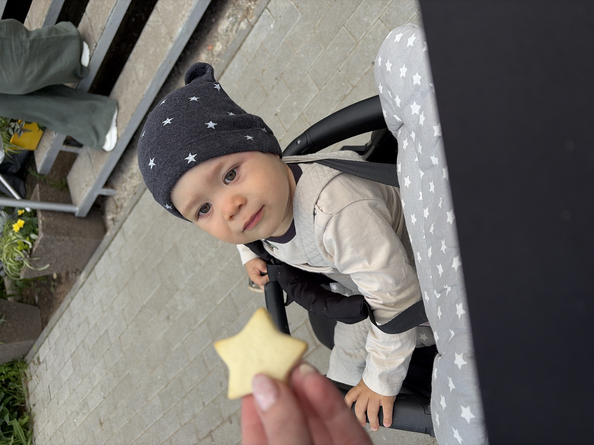 A baby wearing a gray hat with white stars sits in a stroller. A hand holds out a star-shaped cookie. The setting appears to be outdoors near a sidewalk and steps. The stroller has gray and white star-patterned fabric. The location is Chaikovsky, Russia.