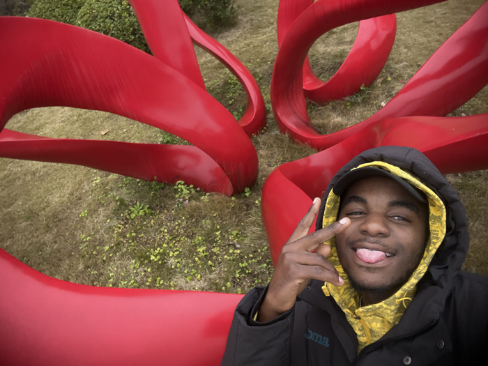 A young man poses for a selfie with a large red abstract sculpture in the background. He wears a black hooded jacket over a yellow patterned hoodie and a baseball cap, flashing a peace sign with his left hand and sticking out his tongue. The sculpture features smooth, curving ribbons of red metal, sitting on a patch of dry grass with small green plants. The photo is taken at eye level, creating a casual and spontaneous feel.