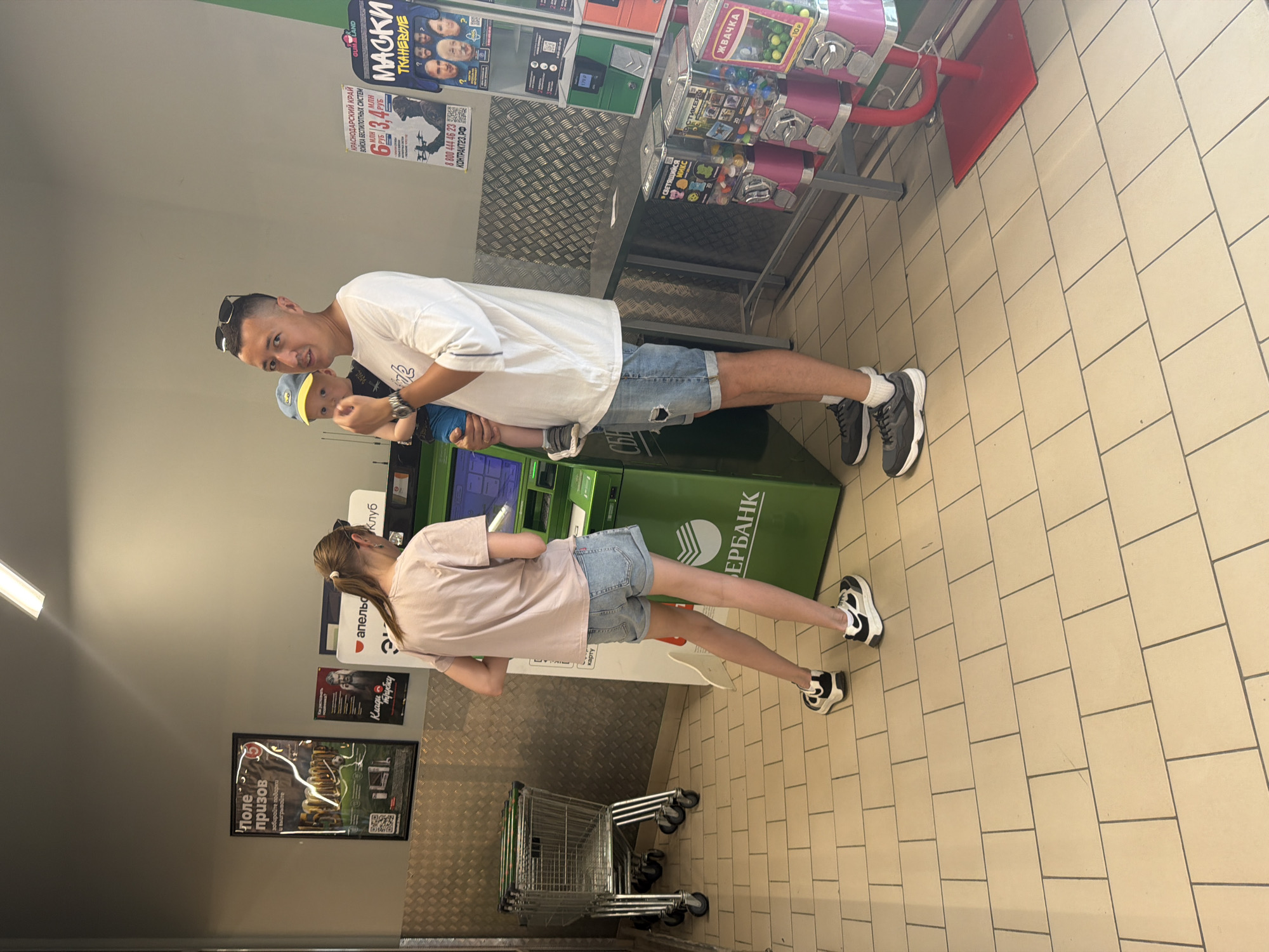 The image depicts an indoor scene, likely a commercial establishment, with a tiled floor and a speckled metallic wall in the background. In the foreground, a man holds a young child. The man is wearing a white t-shirt, denim shorts, and athletic shoes. The child is wearing a blue shirt and a yellow and blue cap. To the left, a young woman in a pink t-shirt and denim shorts stands near a green ATM machine. Several shopping carts are visible in the background to the left. Several posters are displayed on the wall, some featuring text and images, and one appears to be a prize advertisement. To the right, there are several gumball machines. The scene suggests a public space, possibly a supermarket or a service area. The text on the ATM and some posters appears to be in Russian. Based on the visual cues, the location is identified as Leningradskaya, Russia.