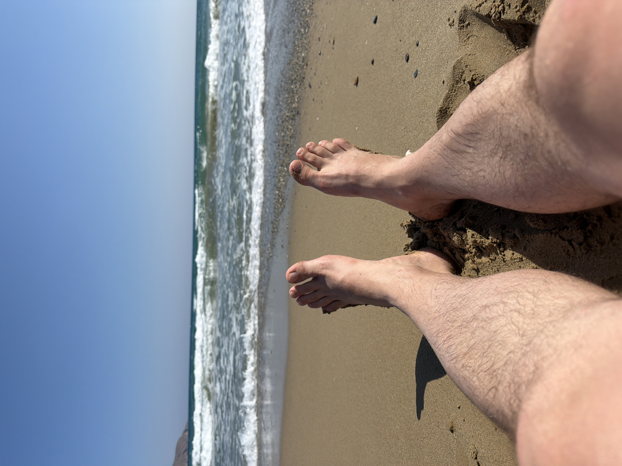 The image captures a close-up, first-person perspective of a person's legs and feet partially submerged in sand on a beach. The sand is light brown and appears damp. The feet are bare, with toes clearly visible and a small amount of sand clinging to them. The legs are hairy and fair-skinned. In the background, the sea is visible with gentle waves breaking on the shore. The water is a turquoise green, and the sky above is a clear, bright blue. A distant landmass, possibly a hill or mountain, is visible on the left horizon. The overall scene suggests a relaxed moment at a beach, likely in Georgioupoli, Greece, given the clear blue sky and turquoise sea.