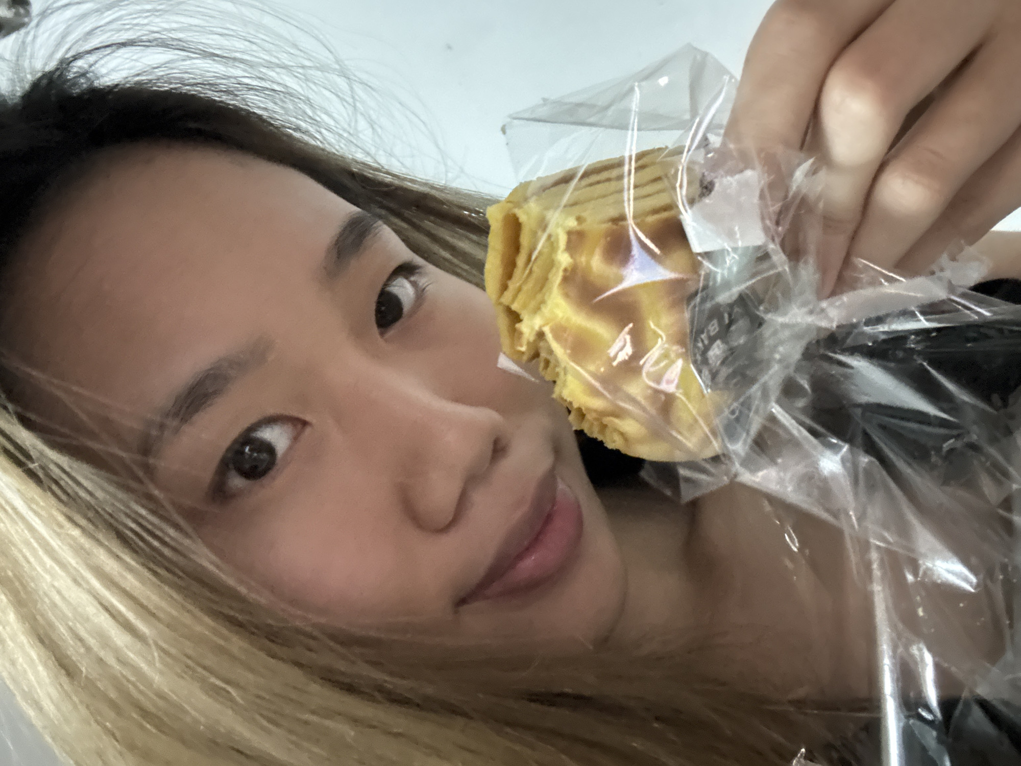 A woman holds a pastry in a plastic wrapper in front of her face. The woman has dark hair with blonde streaks, light skin, and dark eyes. The pastry is a layered yellow color. The background appears to be an indoor setting. The location cannot be determined from the visual information.