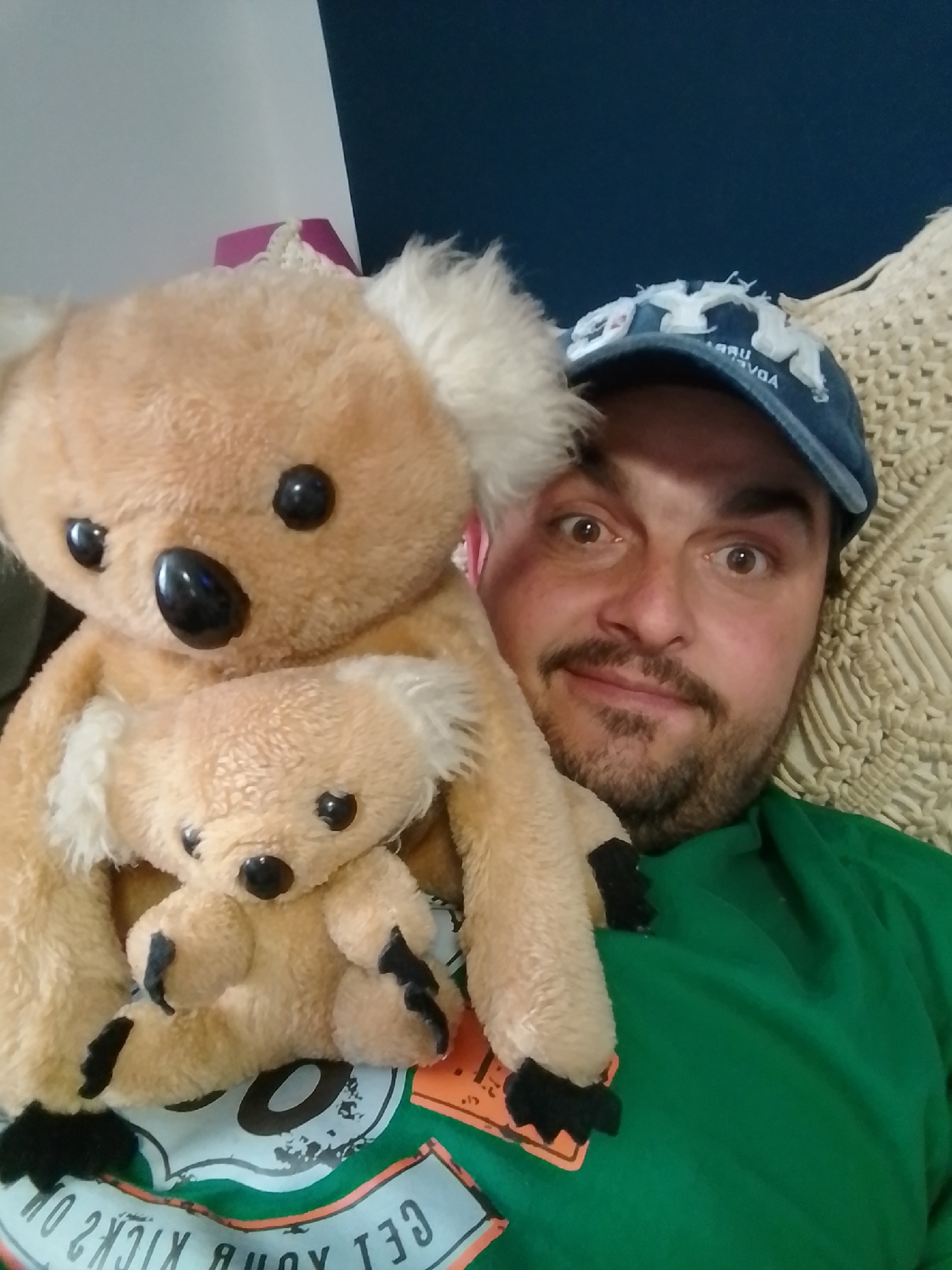 A man wearing a blue baseball cap and a green t-shirt is holding two stuffed koala bears. The larger koala is on the left, and a smaller koala is nested in its arms. The man is looking at the camera with a surprised expression, his eyes wide open. The background appears to be an indoor setting with a dark blue wall and a cream-colored woven texture. The t-shirt has a graphic design on the lower part of the image which is partially visible. This scene is not identifiable as being in the city of Wedel, Germany.