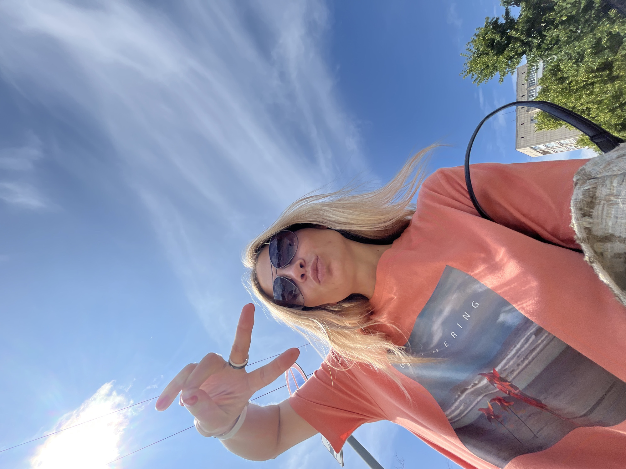 A young blonde woman wearing sunglasses and an orange t-shirt with a picture on it is taking a selfie outdoors. She is making a peace sign with her left hand, and puckering her lips. The sky is blue with some wispy clouds, and the sun is shining brightly, causing some lens flare. In the background, there are green trees, a tall building, and some utility poles with wires. The woman appears to be in the city of Chaikovsky, Russia, given the context provided.