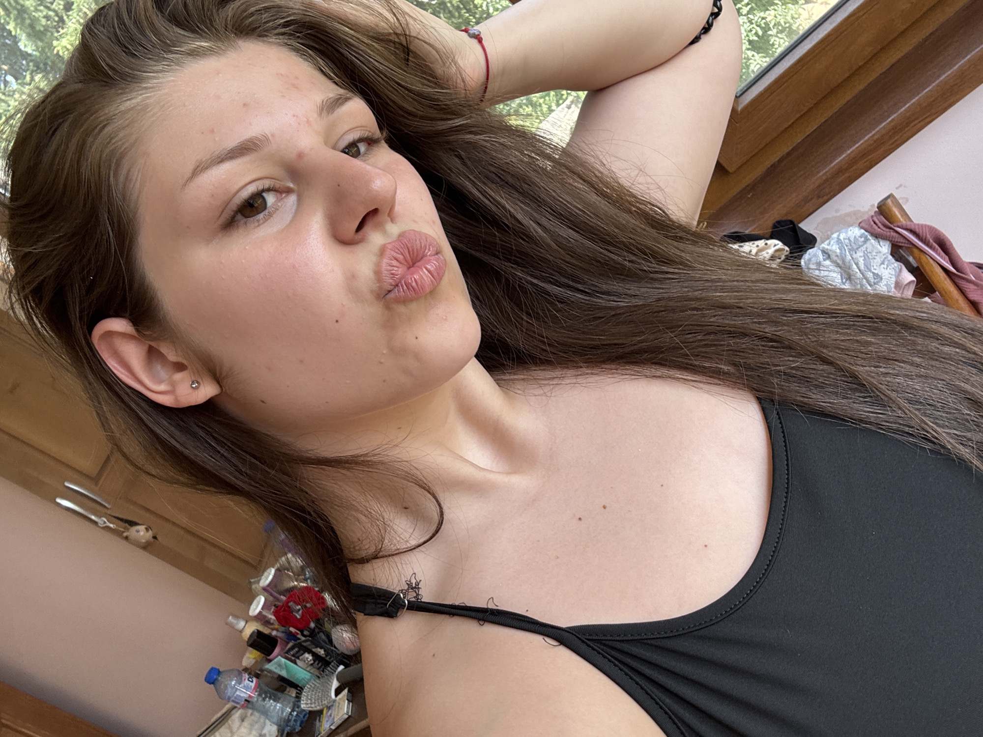 A young woman with long, brown hair is shown in a selfie, puckering her lips. She is wearing a black tank top and has a small stud earring in her left ear. Her right arm is resting on the side of her head. The background includes a dresser with various items on it, such as bottles and toiletries. A window with greenery visible outside is also in the frame. The setting appears to be an indoor room, possibly a bedroom, in Prodanovtsi, Bulgaria.