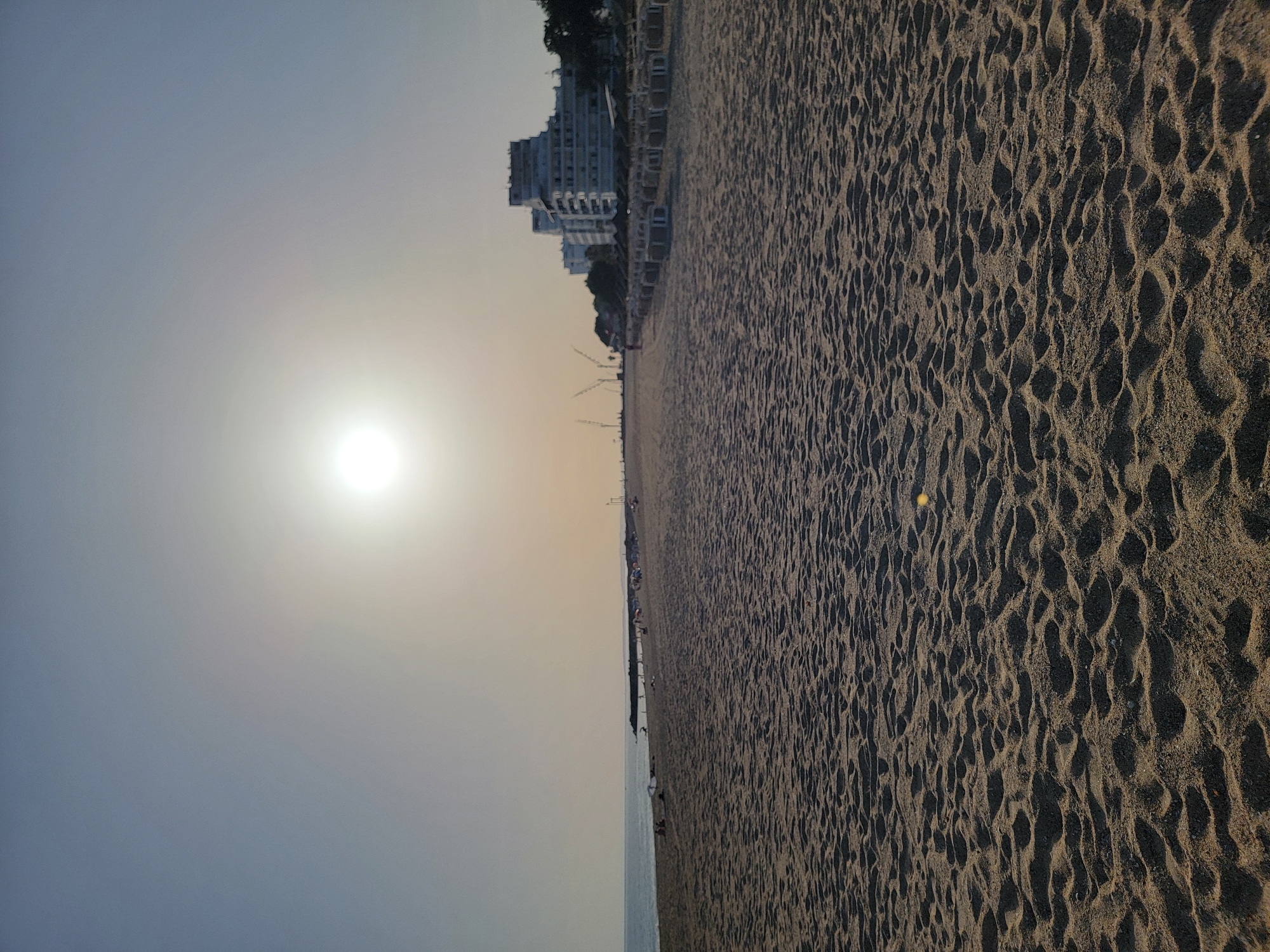 The image depicts a sandy beach in Quarteira, Portugal, under a hazy, bright sun. The foreground is dominated by close-up detail of the rippled sand. In the mid-ground, a few people are visible on the beach, some under umbrellas. Further in the distance, along the coastline, are several buildings, including a multi-story hotel complex. A breakwater extends into the sea on the left side of the image, and construction cranes are visible in the far distance. Beach chairs and umbrellas are neatly arranged in rows along the edge of the beach, suggesting a developed tourist area. The sea is calm and meets the horizon under a pale, bright sky.
