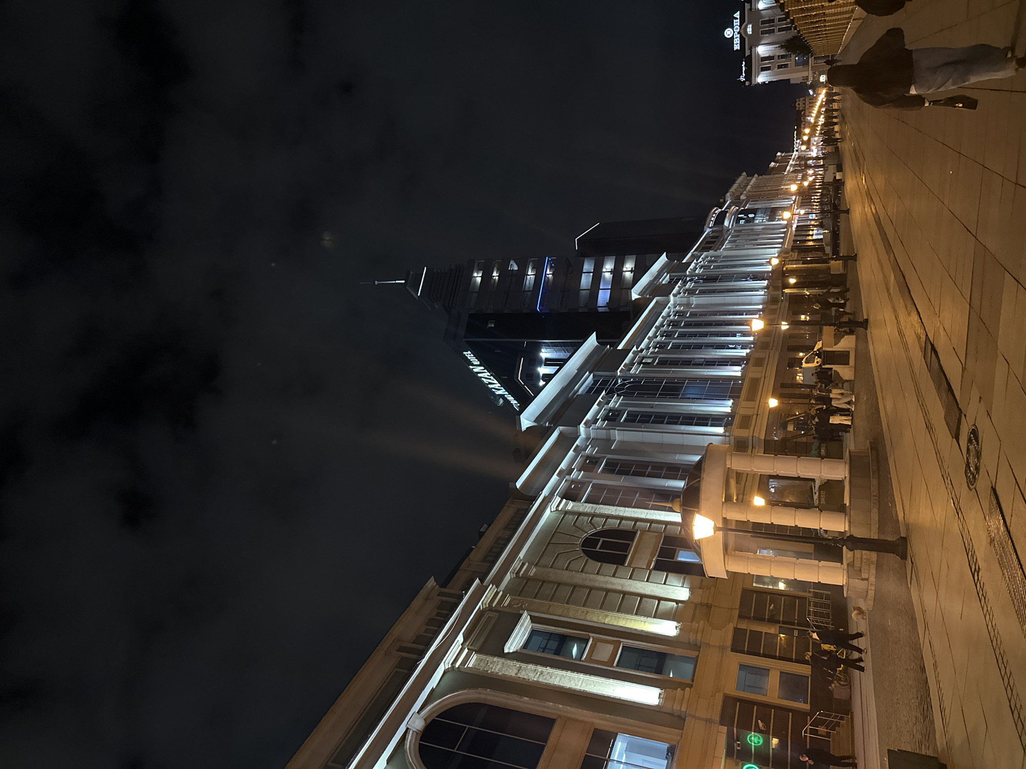 The image shows a nighttime street scene in Kazan, Russia. The street is lined with ornate buildings illuminated by streetlights. In the background, a tall, modern building, labeled "KAZAN RIVIERA HOTEL" is visible, with light beams shooting from it. People are walking along the sidewalk. The sky is dark with some clouds. The perspective is from a low angle, looking up at the buildings. The image is rotated counter-clockwise by 90 degrees.