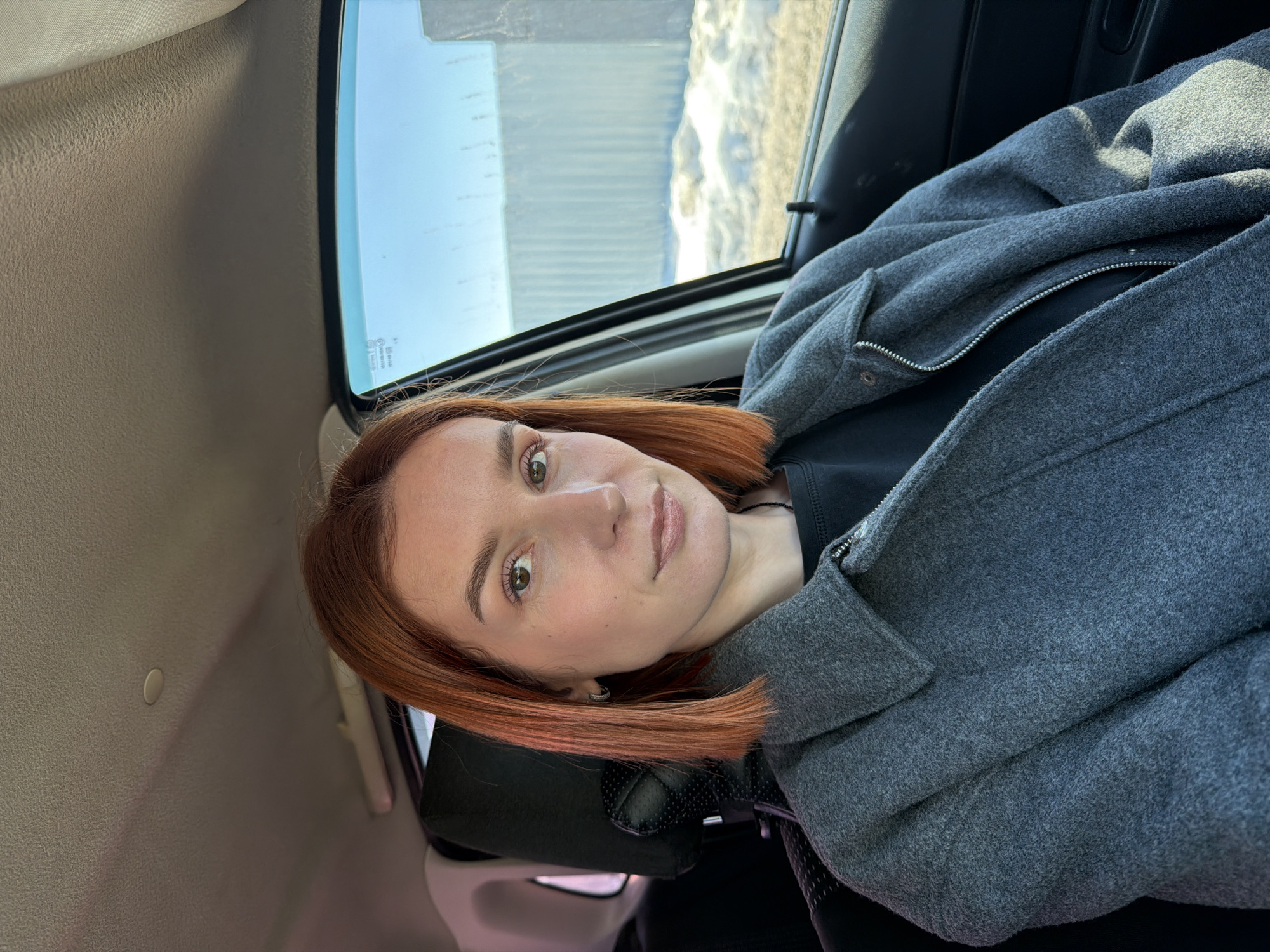 A person with auburn hair, wearing a gray coat, is seated in a vehicle. The exterior environment visible through the window appears to be a landscape, possibly snowy. The location mentioned is Chaikovsky, Russia.