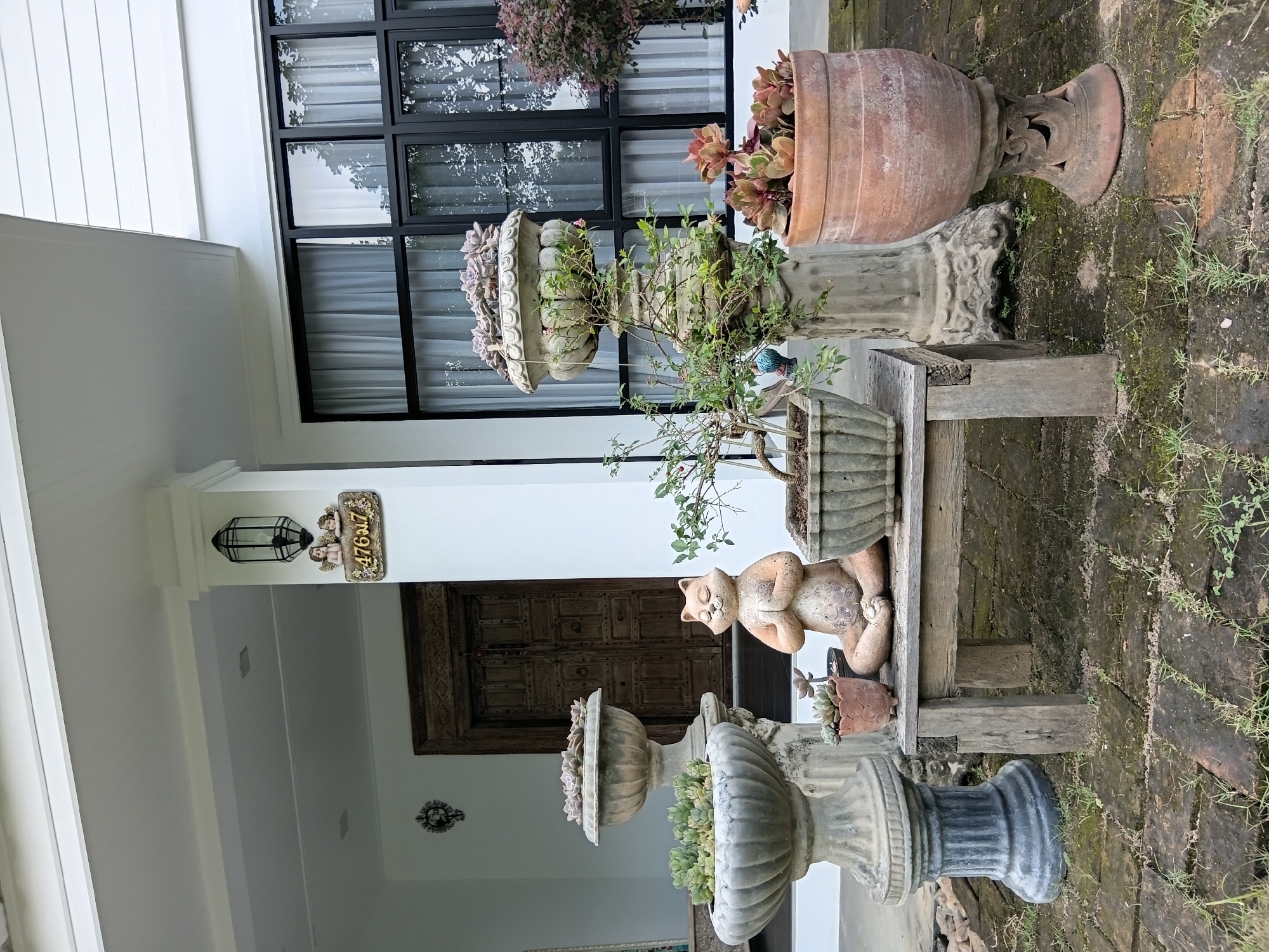 This is a forensic, matter-of-fact description of a scene located in Ban Tong, Thailand. The image depicts the front porch or entryway of a residence. Dominating the foreground are several decorative elements. A light-colored, weathered wooden bench is centrally positioned, supporting a potted plant. To the left of the bench are two large, urn-shaped planters on pedestals, both containing succulents. The planter on the left has a grey, weathered finish, while the one behind it appears to be made of stone and is also filled with succulents. To the right of the bench, a large, terracotta-colored pot on a decorative pedestal holds more succulents. In front of the bench and slightly to the right is a ceramic statue of a cat in a meditative pose, also light brown in color. Behind the bench and planters, a tall, ornate wooden double door serves as the entrance to the house. To the right of the door, a large window with black frames and sheer white curtains is visible. Above the door, a black lantern is mounted on a white pillar, and below it, a decorative house number sign featuring two cherubs and Thai numerals "476 ม.7" is attached to the pillar. The ground is paved with dark, rectangular bricks, some of which are covered in moss and small patches of grass. The overall impression is one of a well-maintained, tastefully decorated home exterior with an Asian influence. There are no people or animals present in the image.