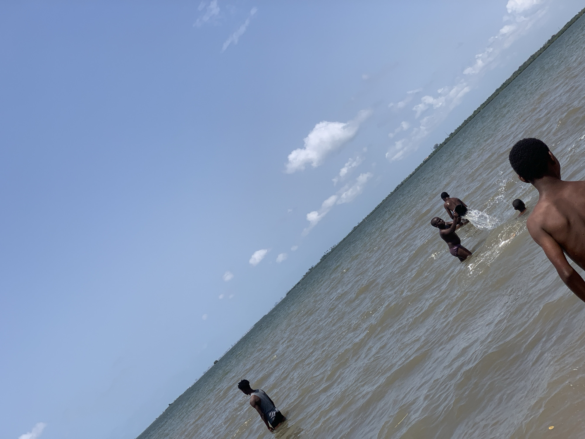 Several people are in the water in Ikorodu, Nigeria. They appear to be swimming and splashing around in the water. The sky is blue and there are some clouds. The water is calm, with small waves. A man is wearing a grey tank top. The picture is taken from the beach or shoreline, looking out towards the water.