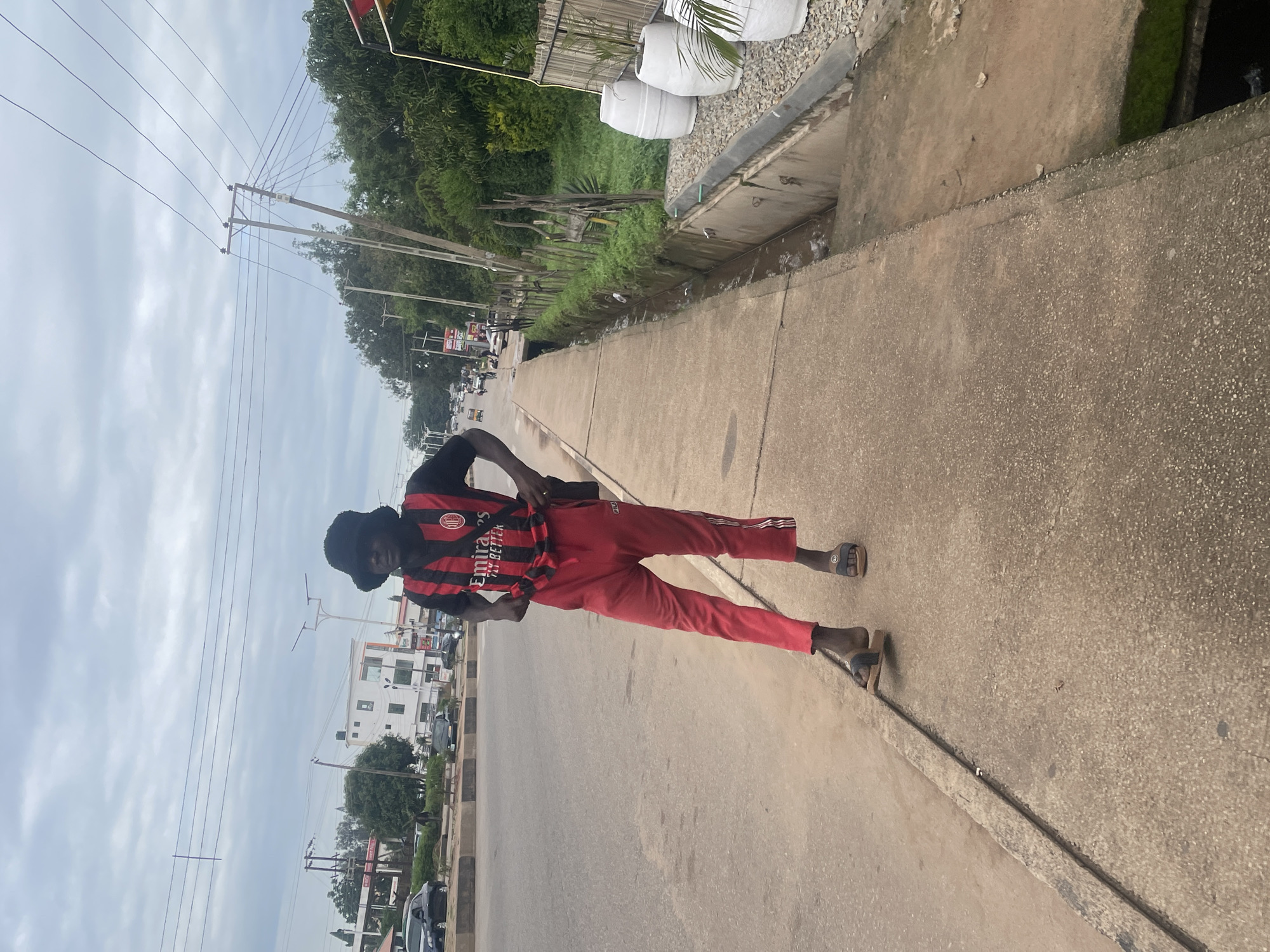 A young male stands on a concrete sidewalk, wearing a black bucket hat, a red and black striped soccer jersey with "Emirates Fly Better" written on it, and red athletic pants. His left foot is positioned over the edge of the sidewalk, and he is holding the hem of his shirt with both hands. He wears brown flip-flops on his feet. The sidewalk is next to a road with a concrete curb. In the background, there is a street with cars, buildings, trees, and utility poles with overhead wires. To the right of the sidewalk, there is a drainage ditch, a wooden fence, and several large white planters. The sky is overcast.