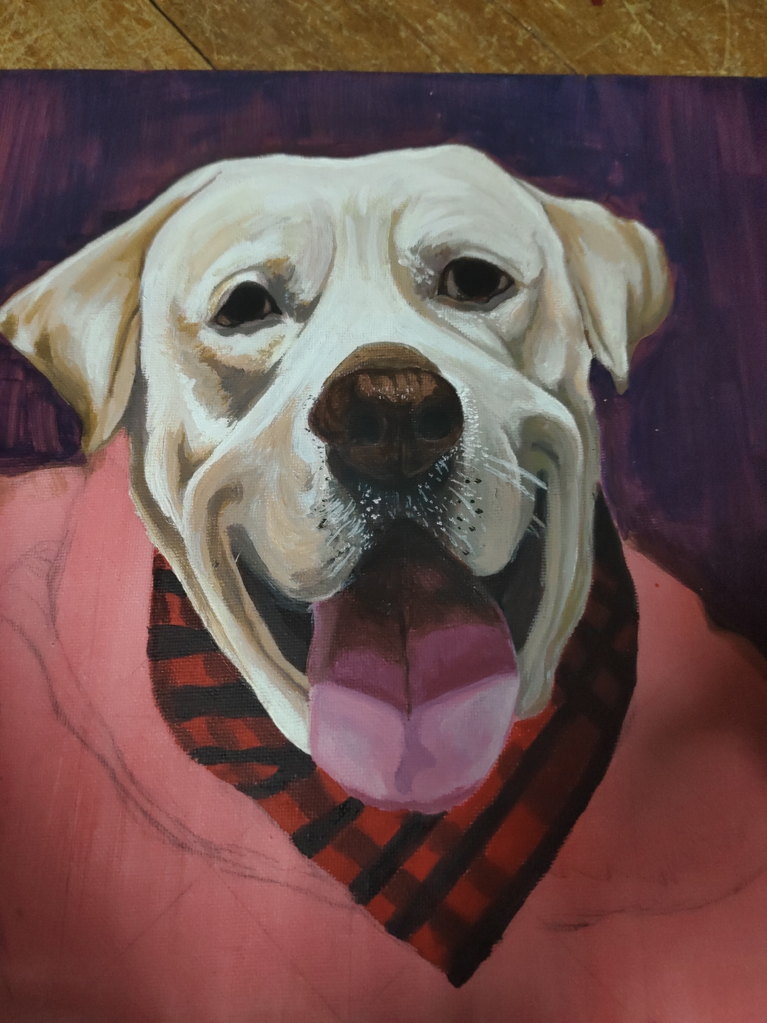The image displays a head-and-shoulders portrait of a light-colored dog, likely a Labrador Retriever, depicted in a painting. The dog's fur is rendered in shades of cream and off-white with visible brushstrokes, indicating texture and contour. Its dark eyes are forward-facing, and a prominent dark brown nose shows subtle highlights and points where whiskers originate. The dog's mouth is open in a wide smile, with a pink tongue extended downward and forward.
Around the dog's neck is a red and black plaid bandana. It is also wearing a pink garment, resembling a shirt or sweater, which covers its chest and shoulders. The pink fabric on the left side of the dog's chest displays faint pencil or charcoal lines, suggesting an unfinished state or preliminary sketching for additional detail.
The background behind the dog's head is a dark purple, characterized by visible brushstrokes. This purple background transitions into the pink of the dog's garment on the lower and side portions of the composition. The overall canvas texture is discernible.
A narrow, light brown, horizontally oriented strip of a textured surface, possibly wood, is visible along the very top edge of the photographic frame, indicating the painting's immediate physical environment. The location of the scene in Davao City, Philippines, is not discernible from the visual content of the image.