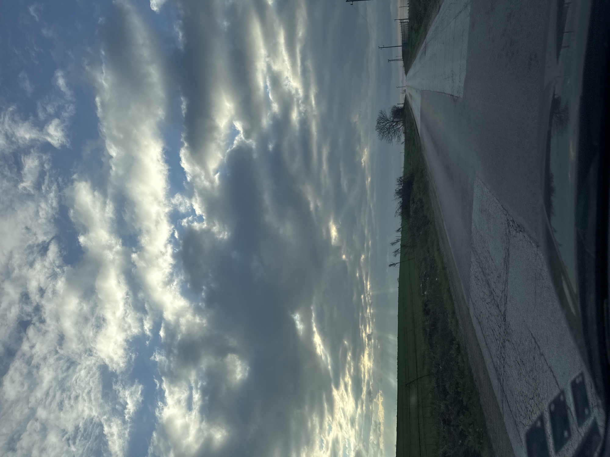 The image shows a rural road in Sofronievo, Bulgaria. The road stretches into the distance, flanked by green fields and sparse vegetation on one side, and the vehicle's interior on the other. Above, the sky is filled with dramatic, striated clouds, with glimpses of blue sky visible. The lighting suggests it is either sunrise or sunset. There are utility poles along the side of the road. The road surface appears to be wet, with some pooling of water. No people are visible.