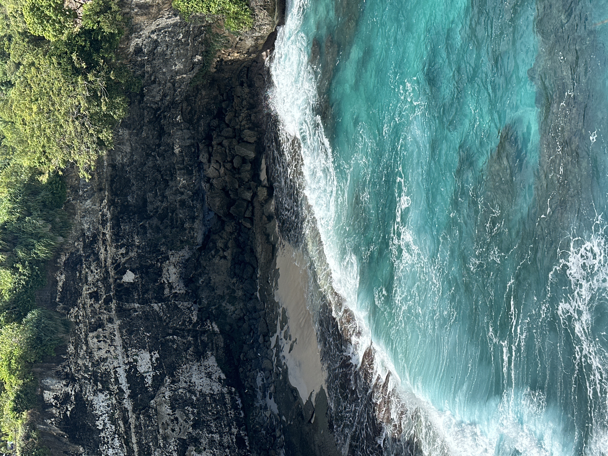 Here is a detailed description of the image:
The image depicts a vertical aerial view of a rugged coastline with vibrant turquoise water. Lush green vegetation grows on the top edge of the dark, rocky cliffs. Waves crash against the shore, creating white foam. The image likely depicts a coastal area in Nusa Penida, Indonesia.