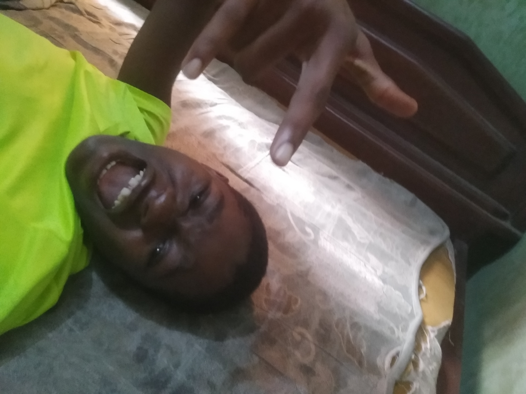 A dark-skinned person with a bright green shirt lies on a bed with a white patterned bedspread, facing the camera. Their mouth is open in a wide expression. Their hand is raised, forming a hand gesture. The bed's headboard is visible, along with part of a wall. The location, Shangisha, Nigeria, is not verifiable based on the provided visual information.