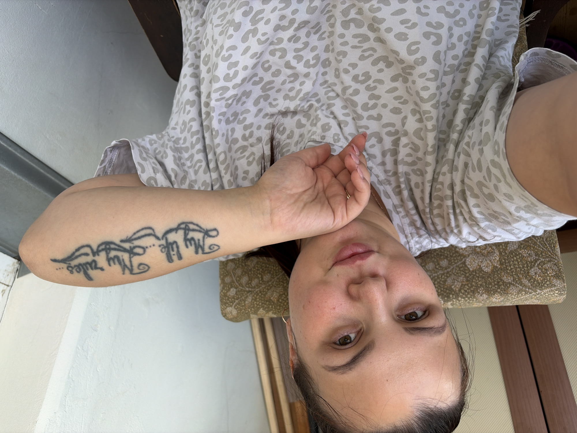 A young woman with dark hair pulled back sits in a chair and takes a selfie. She is wearing a light-colored t-shirt with a grey leopard print. Her left arm is visible, and she has a tattoo on her forearm that reads "My life... My rules...". She is resting her chin on her hand, and her nails are painted a light pink. The background is out of focus, but appears to be an indoor setting with wooden furniture and a textured wall. The scene is not recognizable as being in Izhevsk, Russia.