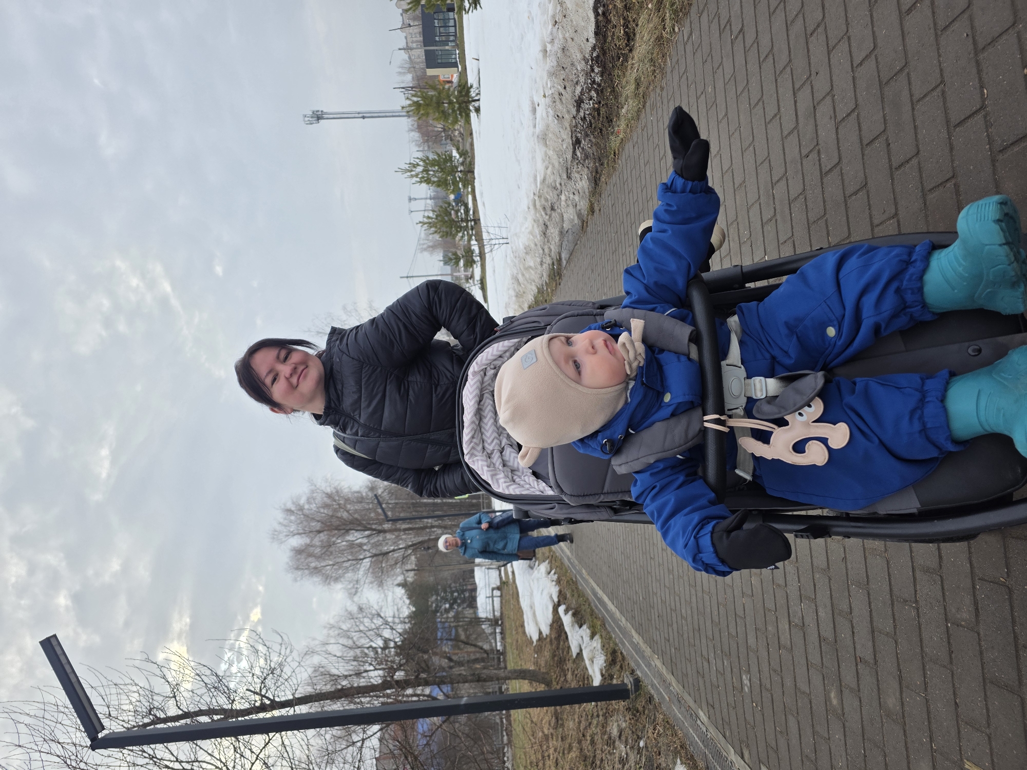 The image captures an outdoor scene in the city of Chaikovsky, Russia, characterized by an overcast, grey sky indicative of late winter or early spring.
In the immediate foreground, an adult female with dark hair, wearing a black quilted jacket, stands behind a grey stroller, facing the camera with a slight smile. Seated in the stroller is a young child, appearing to be an infant or toddler, dressed in a beige beanie with earflaps, a blue snowsuit, black mittens, and turquoise boots. The child's head is turned to the right, and its right hand, covered by a black mitten, is extended forward. A light brown, animal-shaped toy is attached to the stroller's harness. A grey patterned blanket is visible inside the stroller.
The subjects are positioned on a paved pathway constructed from rectangular grey pavers. Flanking the path, patches of melting snow are visible on areas of brown earth and sparse, light-colored vegetation. Bare deciduous trees line the left side of the path and are scattered in the mid-ground, while several small evergreen trees are present on the right.
In the background, an older adult female wearing a light-colored cap, a teal jacket, and dark pants walks away from the camera on the same path, carrying a dark shoulder bag on her left side. Urban infrastructure is evident, including a black, modern streetlight on the left side of the frame and a tall, grey lattice cellular communication tower visible in the distant background on the right. Distant buildings are also partially visible through the trees.