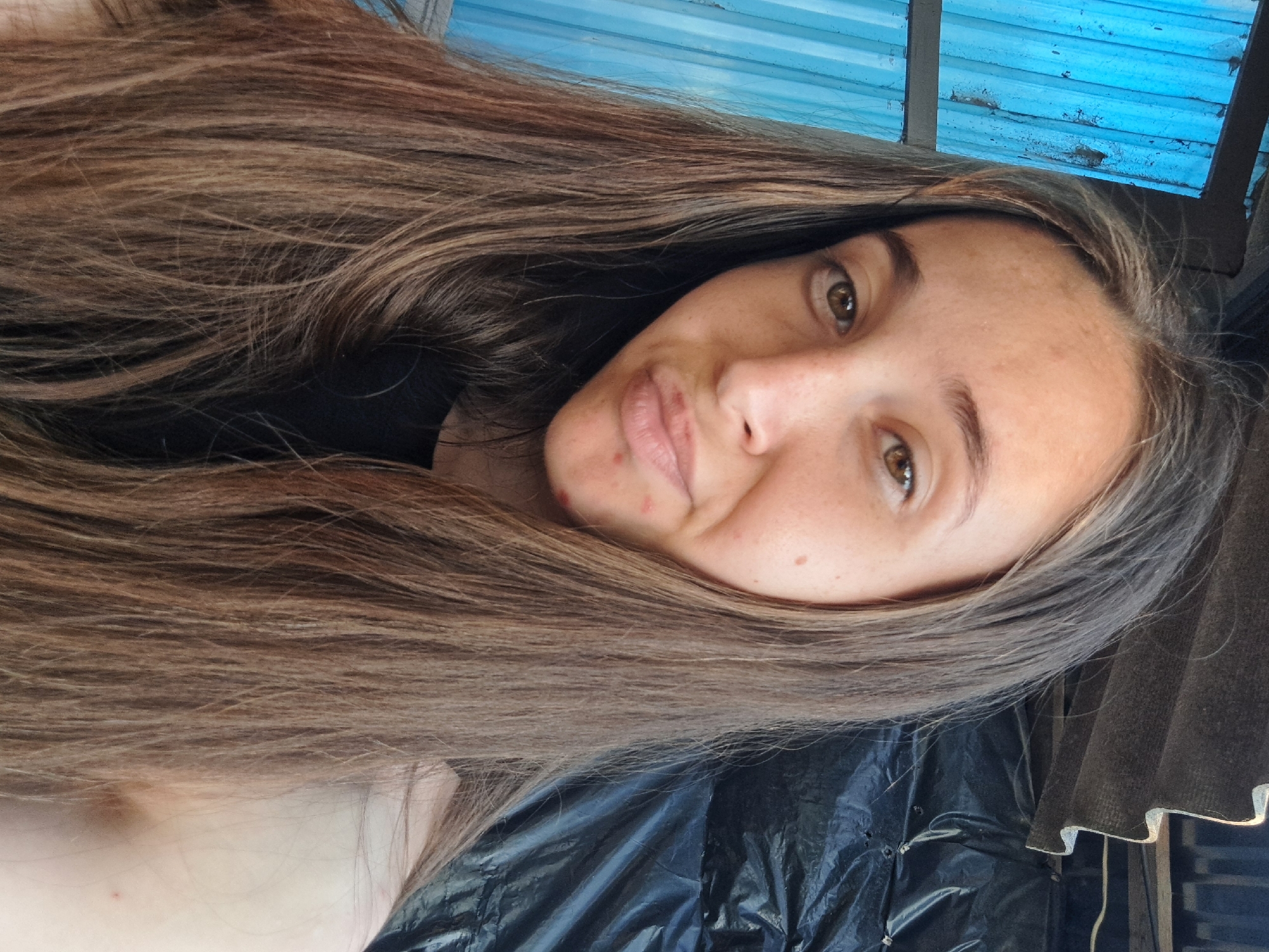 A close-up, selfie-style photo shows a young woman with long, light brown hair and brown eyes. She has a light complexion with freckles and a few blemishes on her face. Her lips are slightly pursed. She is wearing a black top. In the background, to the left, is a blue corrugated metal wall. To the right, there is a dark, crinkled plastic sheeting and a corrugated roof with a jagged edge. The lighting suggests an outdoor setting, possibly during the day. The location is Vypasne, Ukraine.