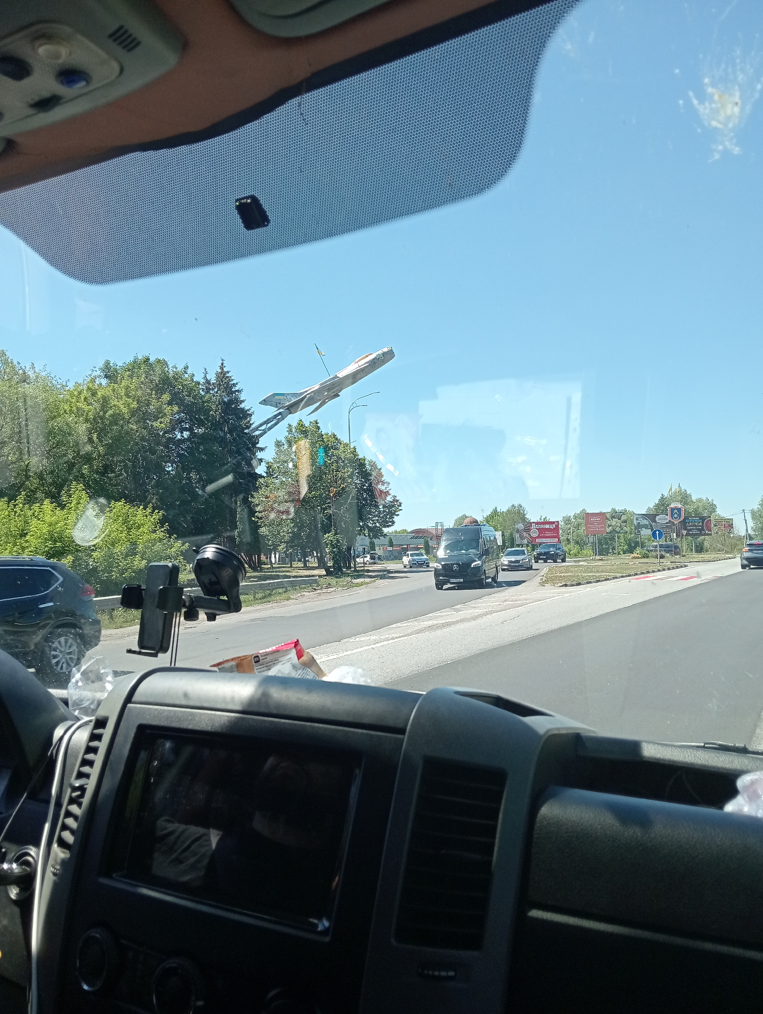 The image is taken from inside a vehicle looking out through the windshield. In the foreground, the dashboard of the vehicle is visible, including a dark screen, air vents, and a black phone mount. Outside the vehicle, a road is visible with several cars and a black van driving on it. There are trees and buildings in the background. The sky is clear and blue. A prominent object in the sky is a silver airplane on a metal pole, appearing to be a monument or art installation. This airplane sculpture is located near the city of Dubno, Ukraine. The overall scene depicts a sunny day on a highway, with the airplane sculpture serving as a distinctive landmark.