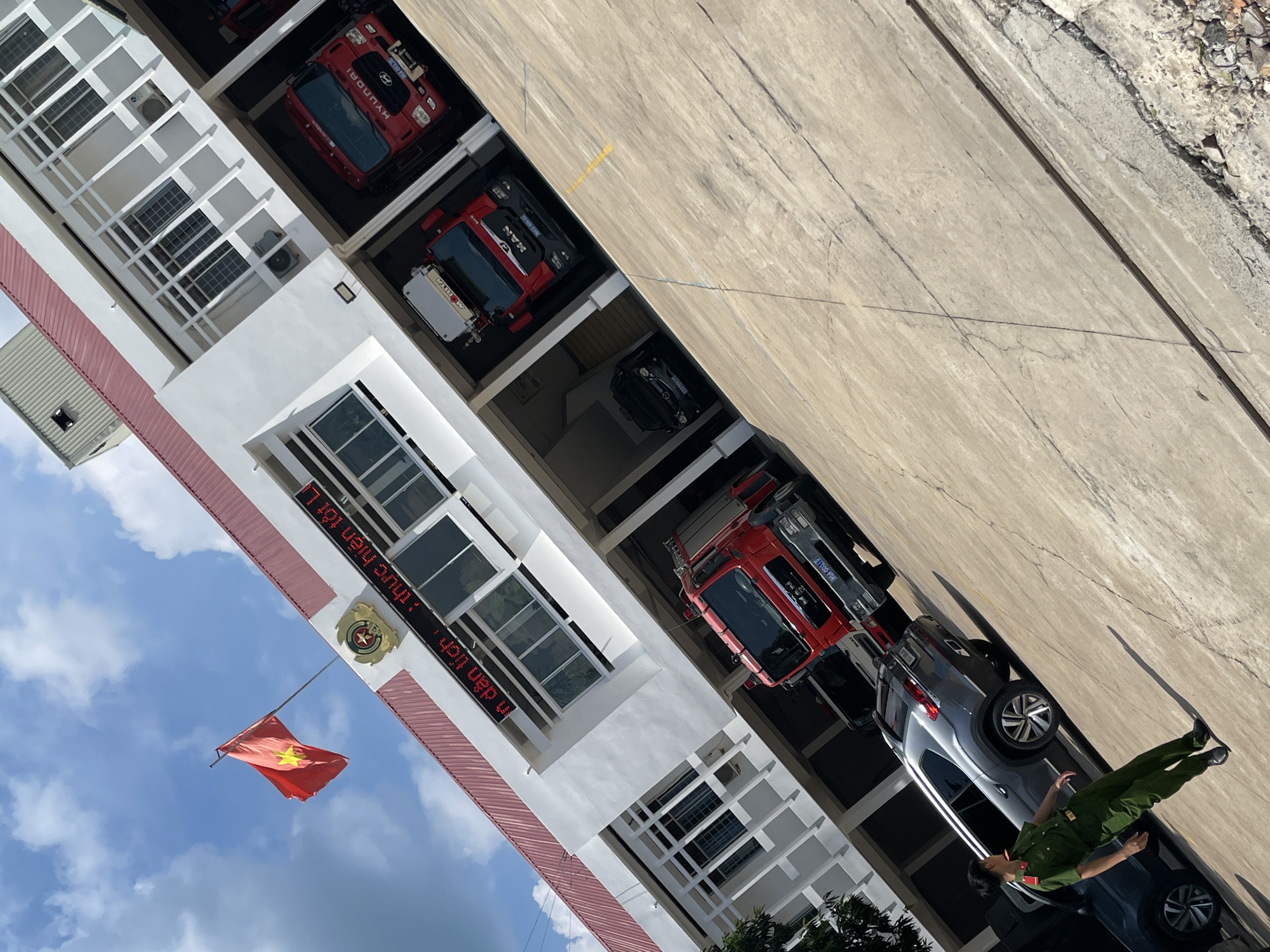 The image depicts the exterior of a fire station in Phu Thuy Ward, Vietnam, on a sunny day. A Vietnamese flag is prominently displayed on a flagpole on the left side of the building, under a bright blue sky with scattered clouds. The fire station is a multi-story white building with a red roof and red trim around the windows. Large garage bays are visible on the ground floor, housing several red fire trucks. One fire truck, a MAN model, is parked in a bay and has its license plate visible: "894 056.17". Another red fire truck, a Hyundai model, is also parked in a bay. A dark-colored SUV is parked in front of the fire station, and a person in a green uniform, possibly a firefighter or police officer, is standing next to it, looking to their left. The ground in front of the building is paved with concrete and shows some wear and tear. The overall scene suggests a functional and well-maintained emergency services facility.
