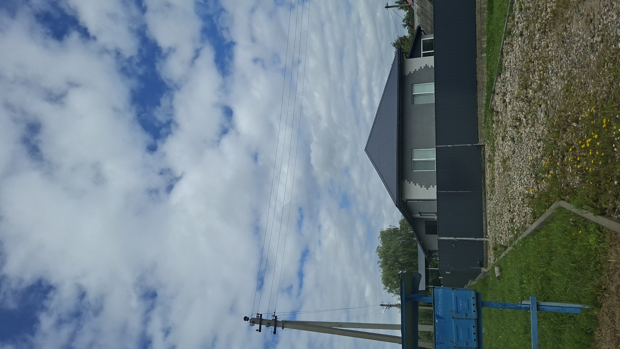 The image depicts a residential property on a cloudy day, likely in the city of Myadzyel, Belarus. The scene is captured from a low angle, focusing on a grey house with a dark metal roof, featuring two prominent windows on the visible facade. A dark grey metal fence encloses the property. To the left of the frame, a blue mailbox stands beside a concrete utility pole with multiple wires. The foreground consists of a gravel driveway bordered by a curb, with patches of green grass and scattered yellow wildflowers. In the background, trees are visible, suggesting a suburban or rural setting. The sky is filled with a mix of white and grey clouds, with patches of blue sky peeking through. No people are present in the image, and no actions or interactions are depicted. The overall impression is a tranquil, everyday scene of a residential area.