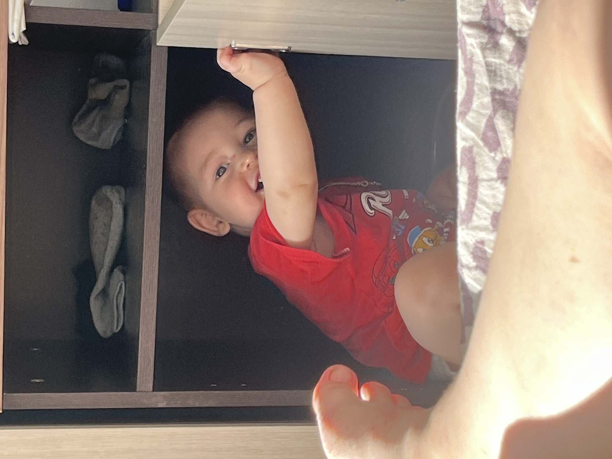 A baby is peeking out of a shelf in a closet, their left arm extended and holding onto the side of the closet door. The baby is wearing a red shirt with a graphic print. The background is dark, with a few items on the shelves above the baby, including what appear to be folded gray socks. The closet door is light colored. In the foreground, a person's leg and foot are visible, bathed in sunlight. The scene is likely indoors, in a residential setting. There is no information available to determine the location in the city of Chelyabinsk, Russia.