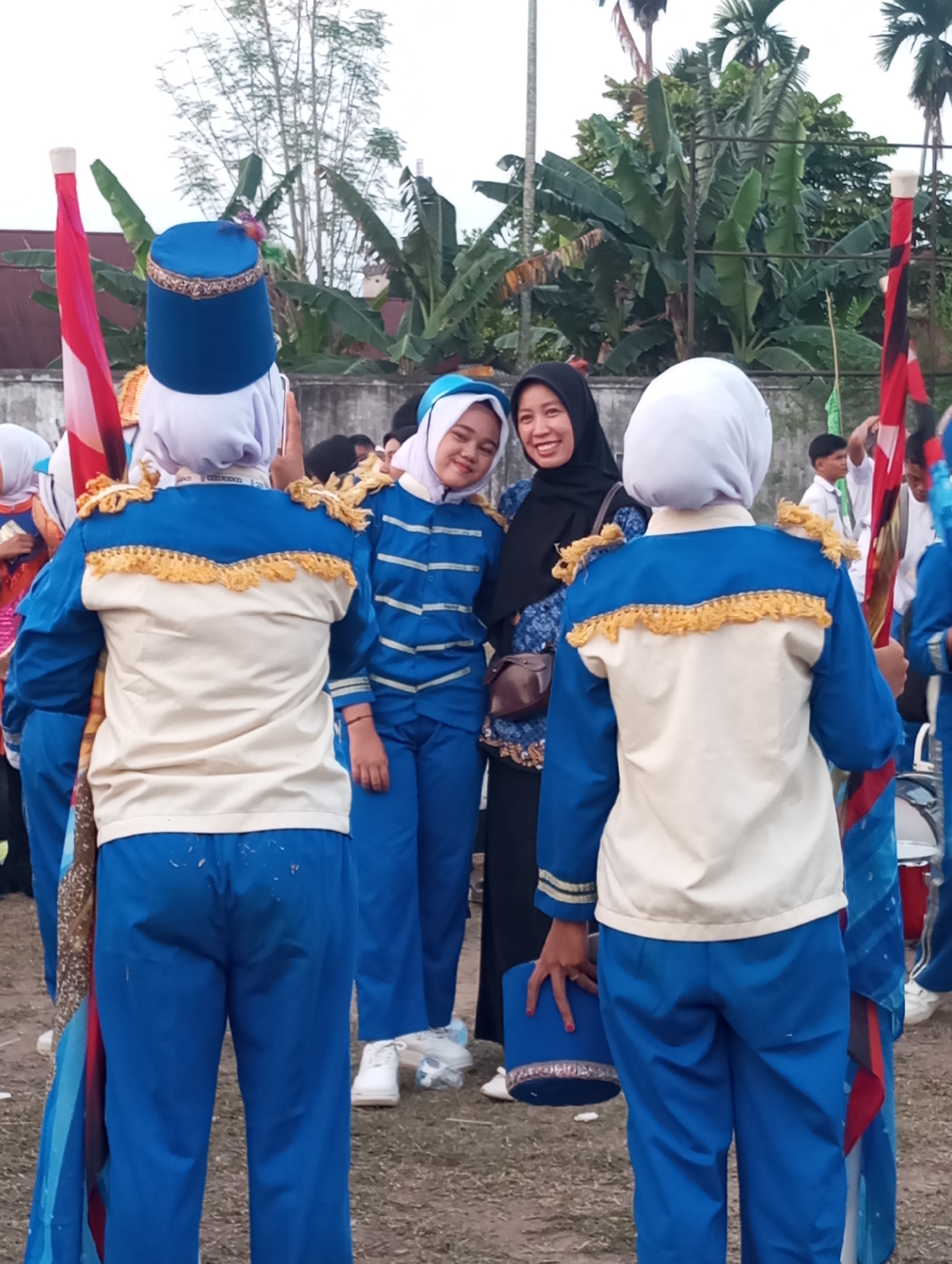 In Candirejo, Indonesia, a group of individuals in marching band uniforms, predominantly blue and cream with gold trim, are gathered outdoors. Two individuals in the foreground wear blue caps and matching uniforms with decorative gold fringe on the shoulders and chest area. Behind them, a woman in a black hijab and patterned blue top smiles at the camera, standing next to a young person also wearing a hijab and a blue and white striped marching band uniform. Other band members in similar attire are visible in the background, along with trees and foliage. A flag with red and white elements is held by one of the uniformed individuals. The ground appears to be dirt or gravel. The scene suggests a public event or practice session for a marching band.