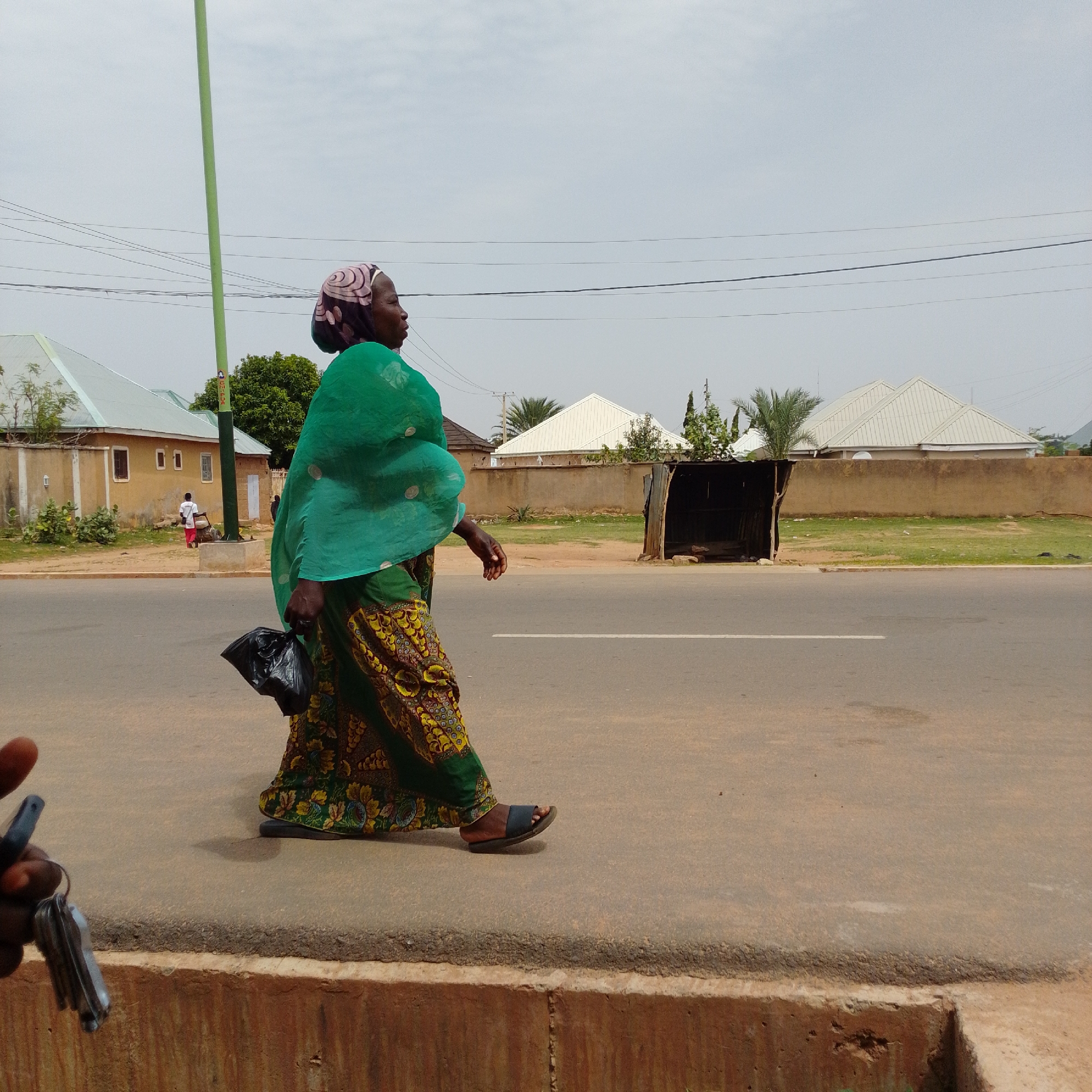 A woman walks on a paved sidewalk next to a road in Gombe, Nigeria. She is wearing a green headscarf and a colorful patterned skirt, and she is carrying a black plastic bag in her left hand. In the background, there are buildings with light-colored roofs, trees, and power lines. The sky is overcast. The sidewalk appears to be made of concrete and is cracked in places. The road is asphalt. A person's hand holding a set of keys is visible in the lower left corner of the image.