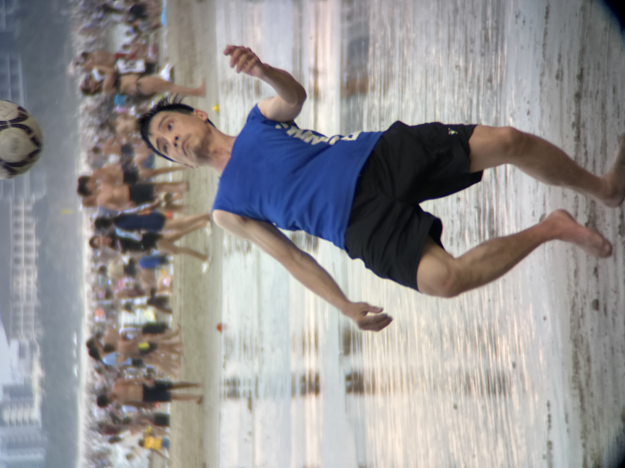 The image shows a crowded beach scene, likely in Da Nang, Vietnam, based on the architecture in the background resembling beachfront resorts common in the city. In the foreground, a man in a blue sleeveless shirt and black shorts is captured mid-action, attempting to head a soccer ball that is out of frame but implied to be above and to his left. He is looking upwards with an intense expression, his body angled as if bracing for impact. The beach is wet, with shallow water reflecting the sky and the distant crowd. Numerous other people are visible in the mid-ground and background, some walking, some standing, and others relaxing. The overall atmosphere suggests a sunny, active day at a popular tourist destination.