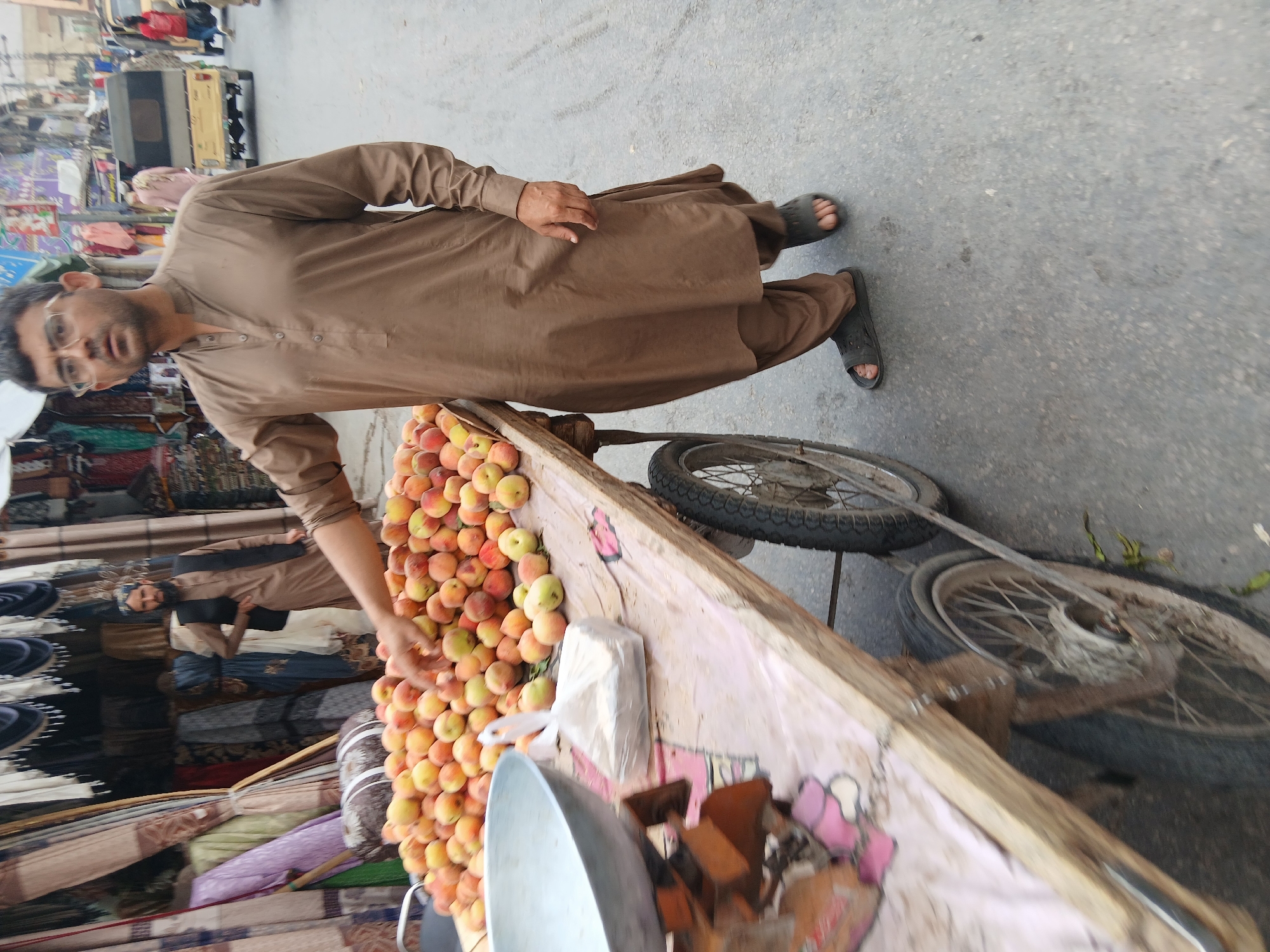 A man stands in the street next to a cart laden with peaches. He has dark hair, wears glasses, and is dressed in a light brown traditional outfit and sandals. The cart has two wheels and is painted with a rough design of pink hearts. The street appears to be in a busy area, likely in Quetta City Tehsil, Pakistan. Other goods are visible stacked beside the cart.