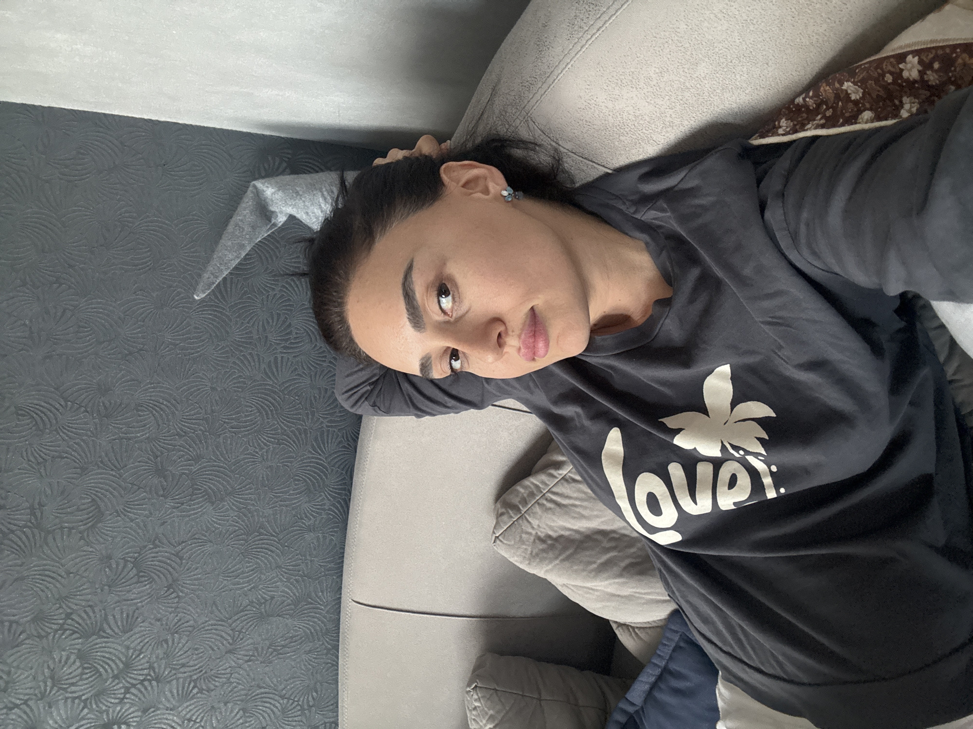 A woman lies on a gray couch. She wears a black shirt with a white "Lover" graphic. She has dark hair pulled back, dark eyebrows, and earrings. The couch is situated against a gray wall with a textured pattern. Pillows are visible on the couch. The location appears to be indoors. No information in the image supports a connection to Tbilisi, Georgia.