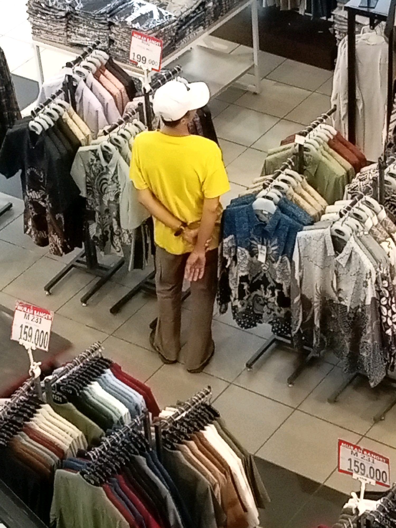 A person wearing a yellow t-shirt and white cap is standing in a retail store, possibly a clothing store, in Batam, Indonesia. The person is facing away from the camera, with their hands behind their back. The store appears to be selling various types of clothing, with racks of shirts and folded garments visible in the background. Several price tags are displayed, with one showing "159.000" and another "199.000". The floor is tiled, and the lighting suggests an indoor environment. The overall scene depicts a typical retail setting with merchandise and a customer.