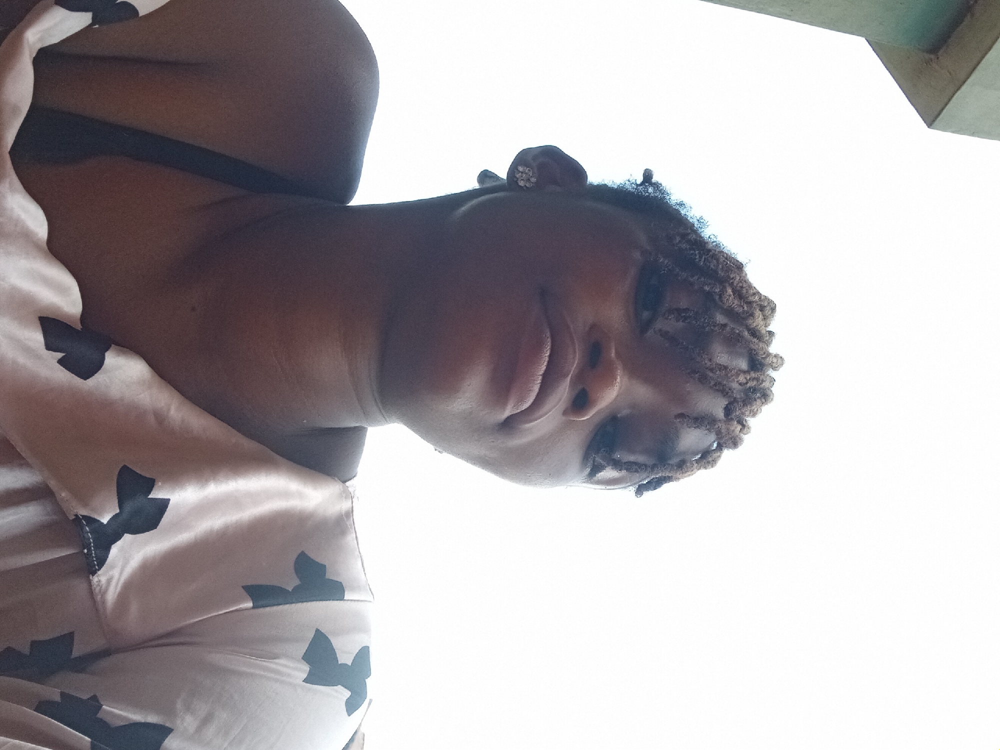 A person with dark skin and braided hair is viewed from below. The person is wearing a tan and brown patterned garment. The location of the photograph is unknown and there is no indication it was taken in Shagamu, Nigeria.