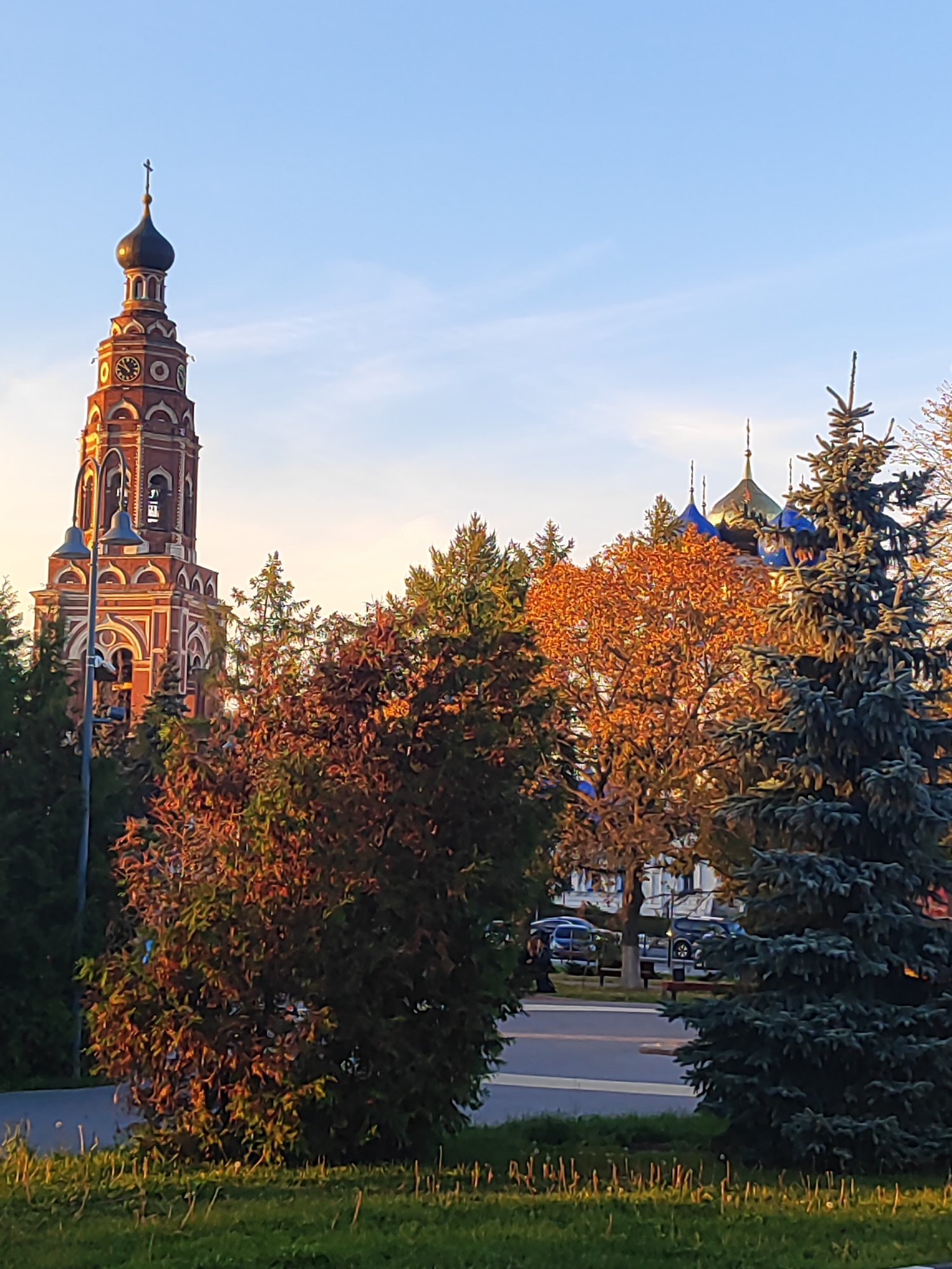The image depicts a view towards the Resurrection Cathedral in Bronnitsy, Russia. The cathedral's tall, red brick bell tower dominates the left side of the frame, adorned with a clock face and topped with a dark, onion-shaped dome and a cross. In the background, other gilded domes and blue roofs of the cathedral complex are visible, partially obscured by trees. The foreground is filled with foliage, including evergreen trees and deciduous trees displaying autumnal colors of orange and red. A paved road with parked cars and a bench is visible in the middle ground, suggesting a public area. The scene is bathed in the warm, golden light of either sunrise or sunset, casting long shadows and highlighting the architectural details and natural elements. The sky is clear blue with subtle clouds.