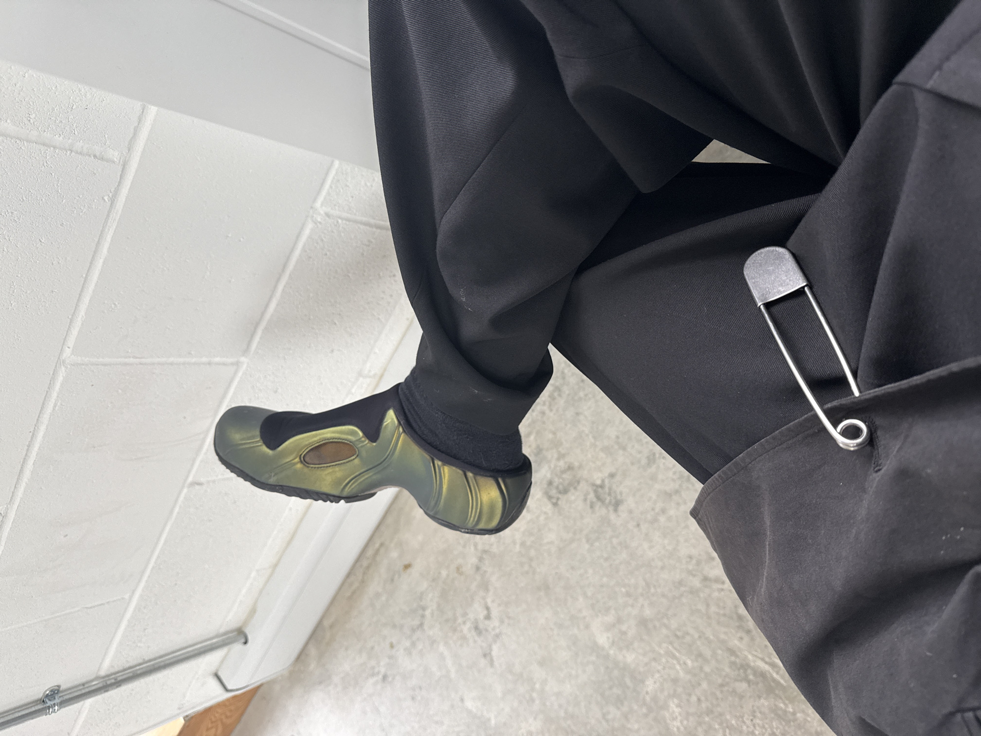 A person is seen from the knees up, seated and wearing black trousers, black socks, and iridescent green and black shoes. A large, silver safety pin is clipped to the pocket of the trousers. The background is a white brick wall and concrete floor. The location is not discernible from the image.
